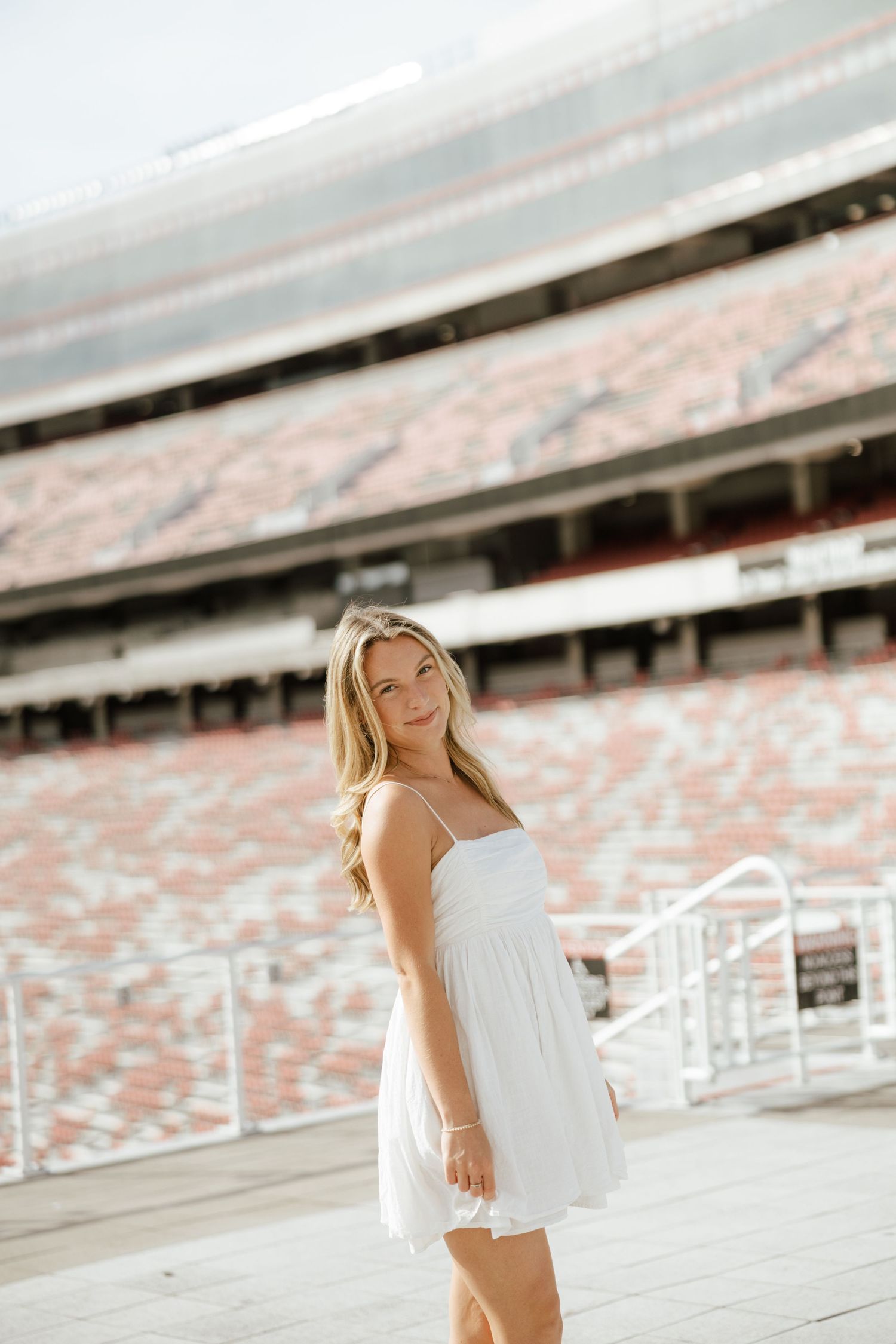 Young blonde woman in white sundress smiling at camera in front of large stadium seating.