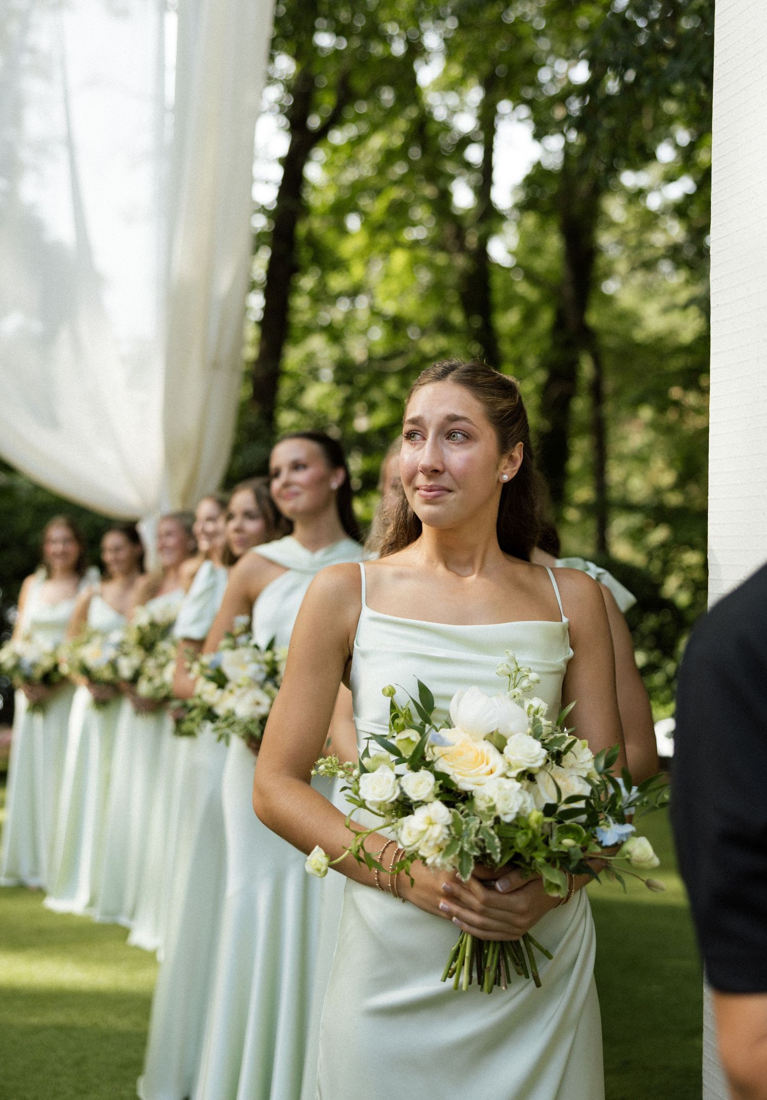 Bridesmaids in mint green gowns holding white floral bouquets stand in a line during an outdoor wedding ceremony.