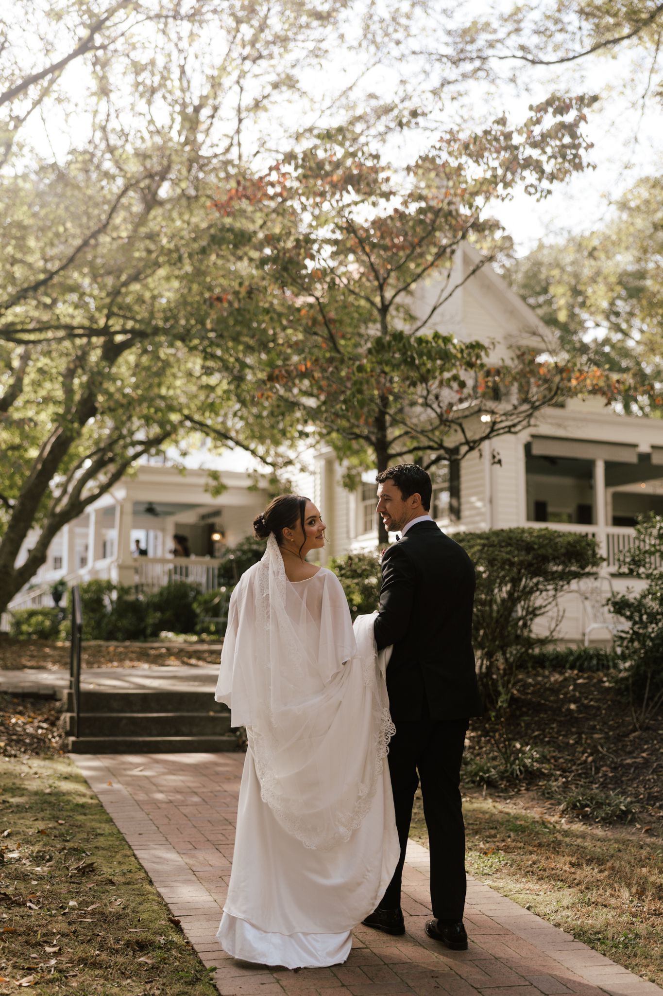 Wedding couple in formal attire stroll along brick pathway surrounded by trees.