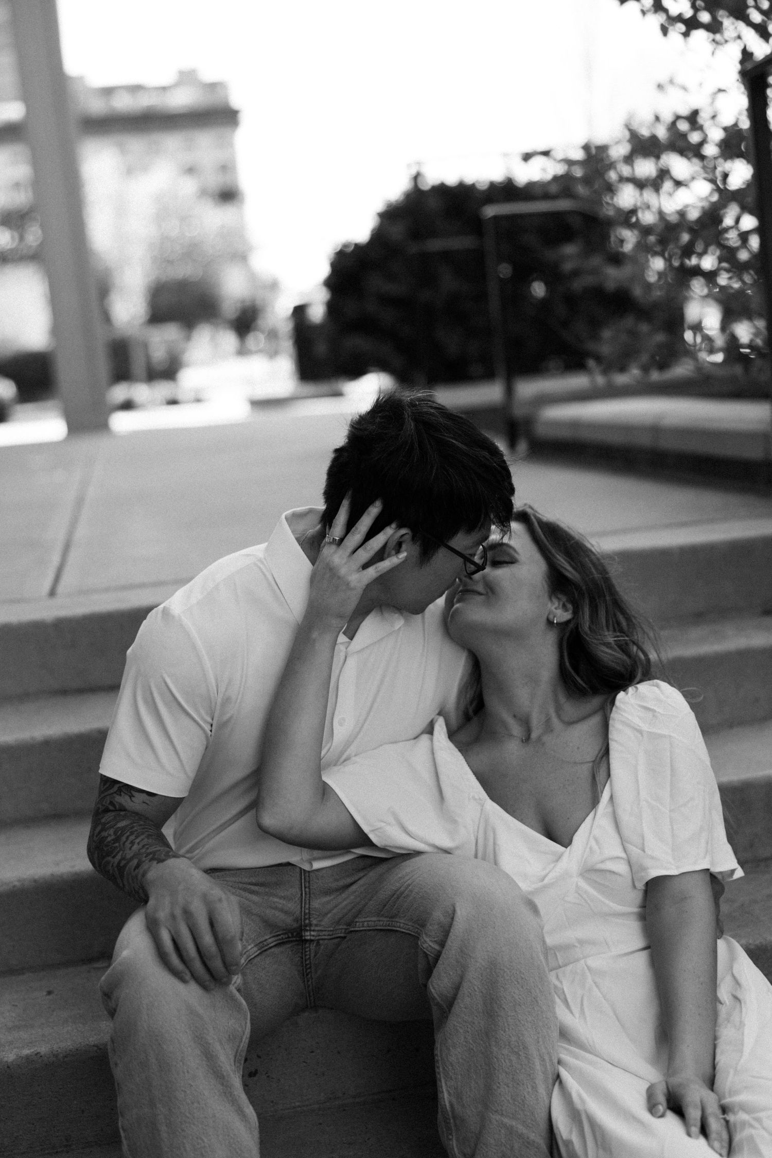 Black and white photo of a romantic couple sharing a kiss on outdoor steps in an urban setting.