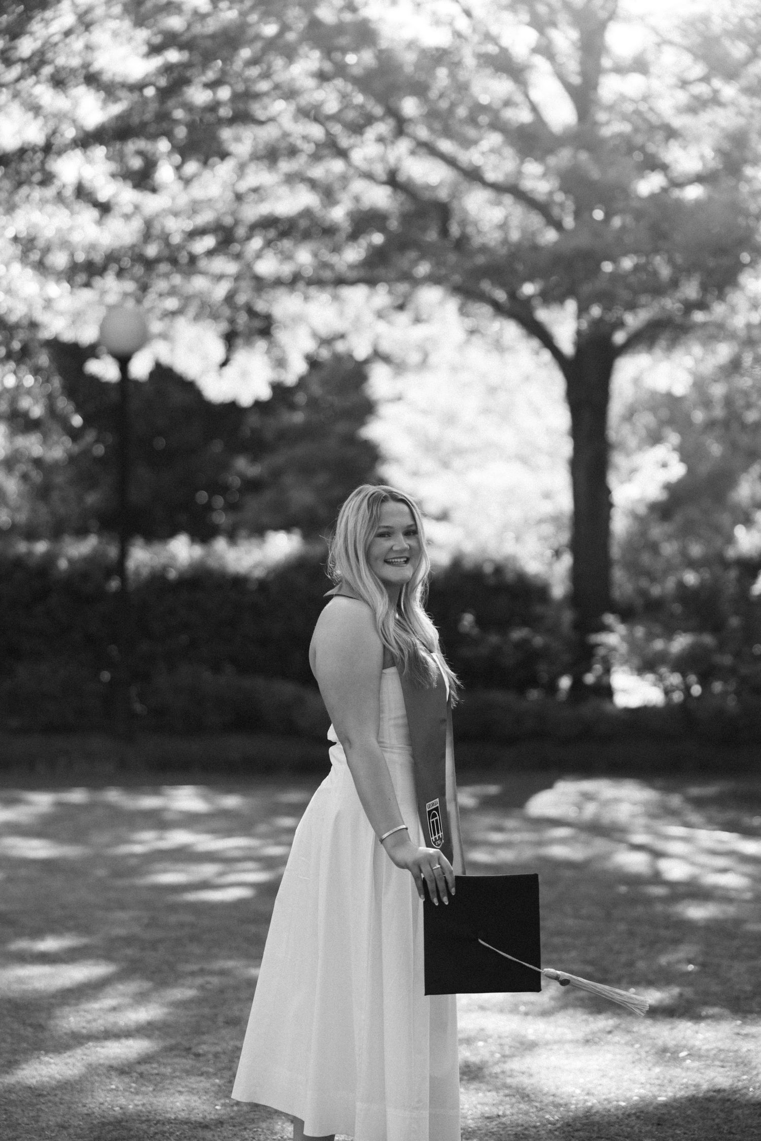 Black and white photo of a smiling blonde woman in a white dress holding a graduation cap outdoors.