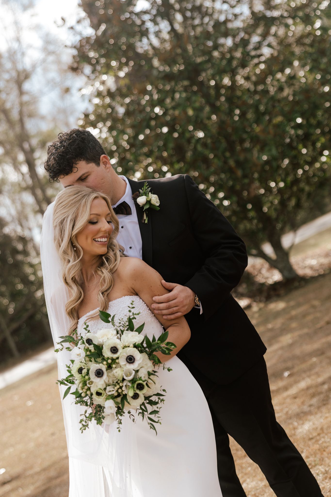 Smiling bride in strapless gown holding white floral bouquet while groom in tuxedo embraces her outdoors.