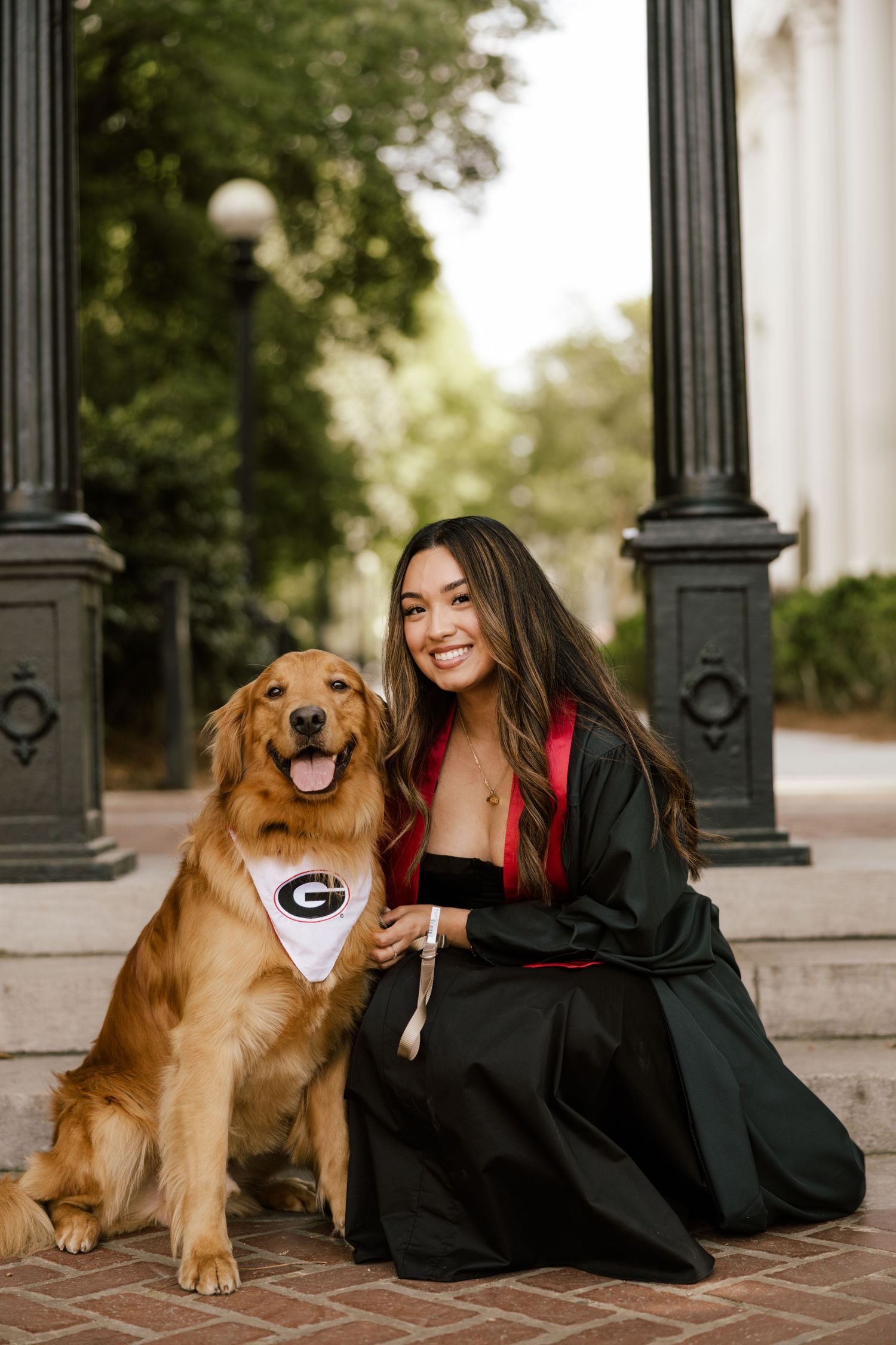 A smiling graduate in black and red gown poses with a golden retriever wearing a University of Georgia bandana.