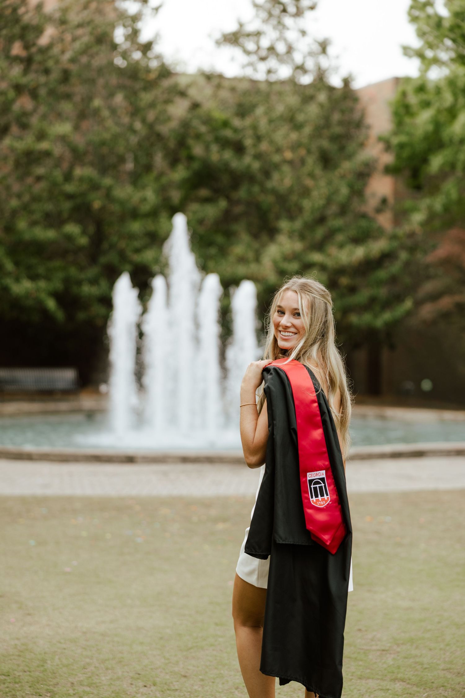 Smiling graduate holding black gown and red stole outdoors near fountain on college campus.