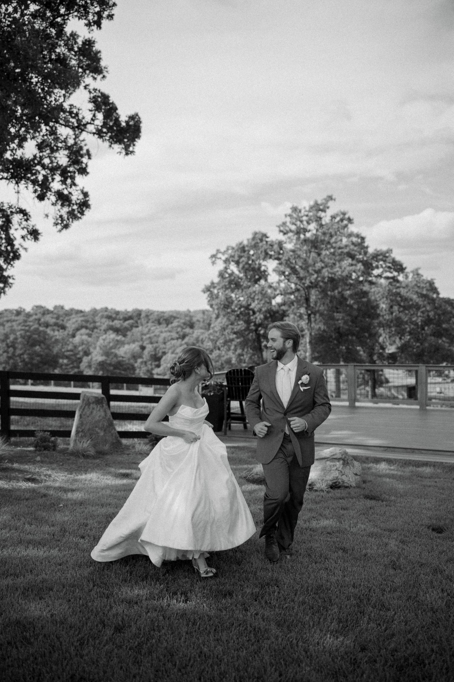 Black and white photo of a bride and groom joyfully running together on a grassy lawn at an outdoor wedding venue.