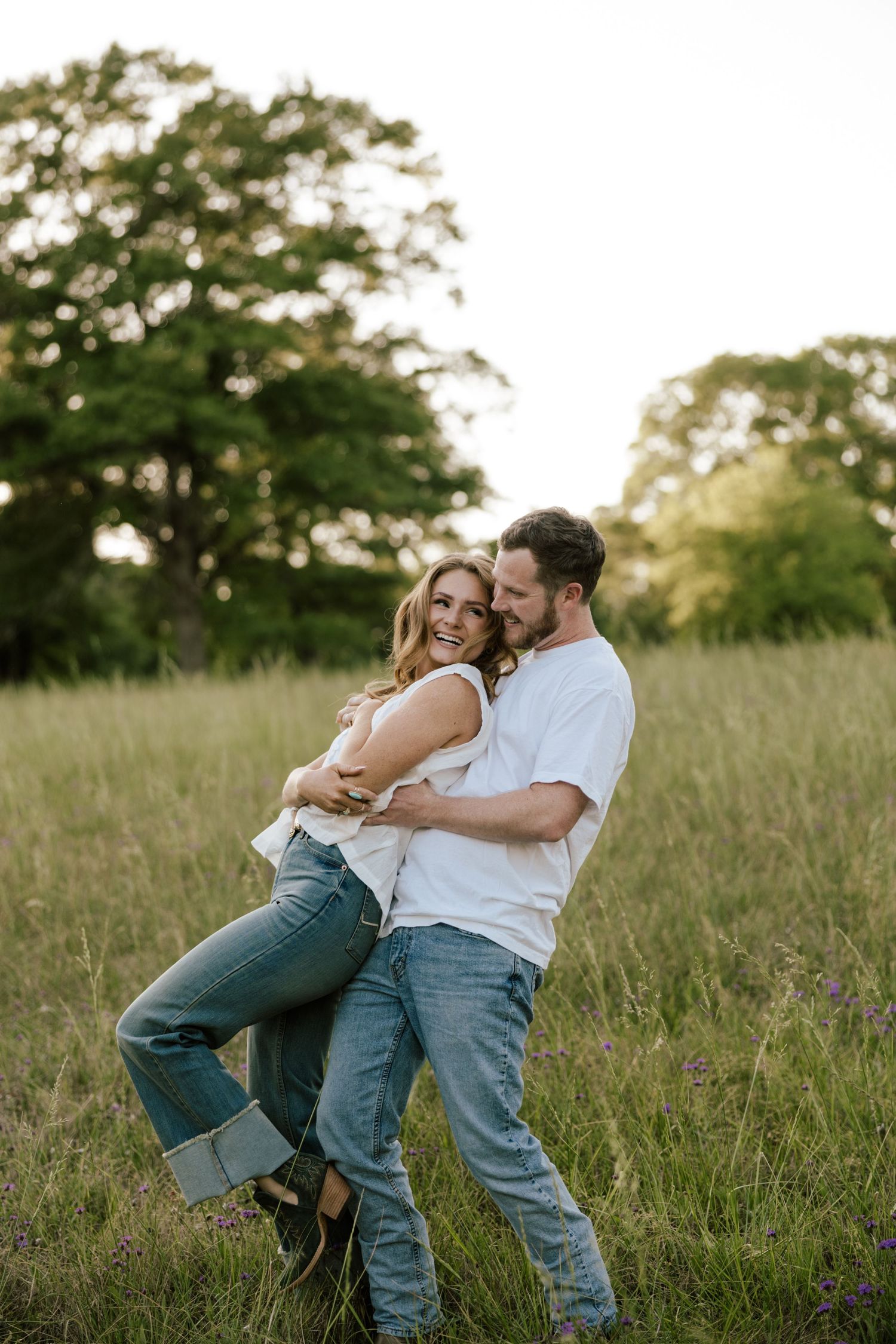 A happy couple embraces and laughs together in a sunlit green meadow, both wearing white tops and jeans.