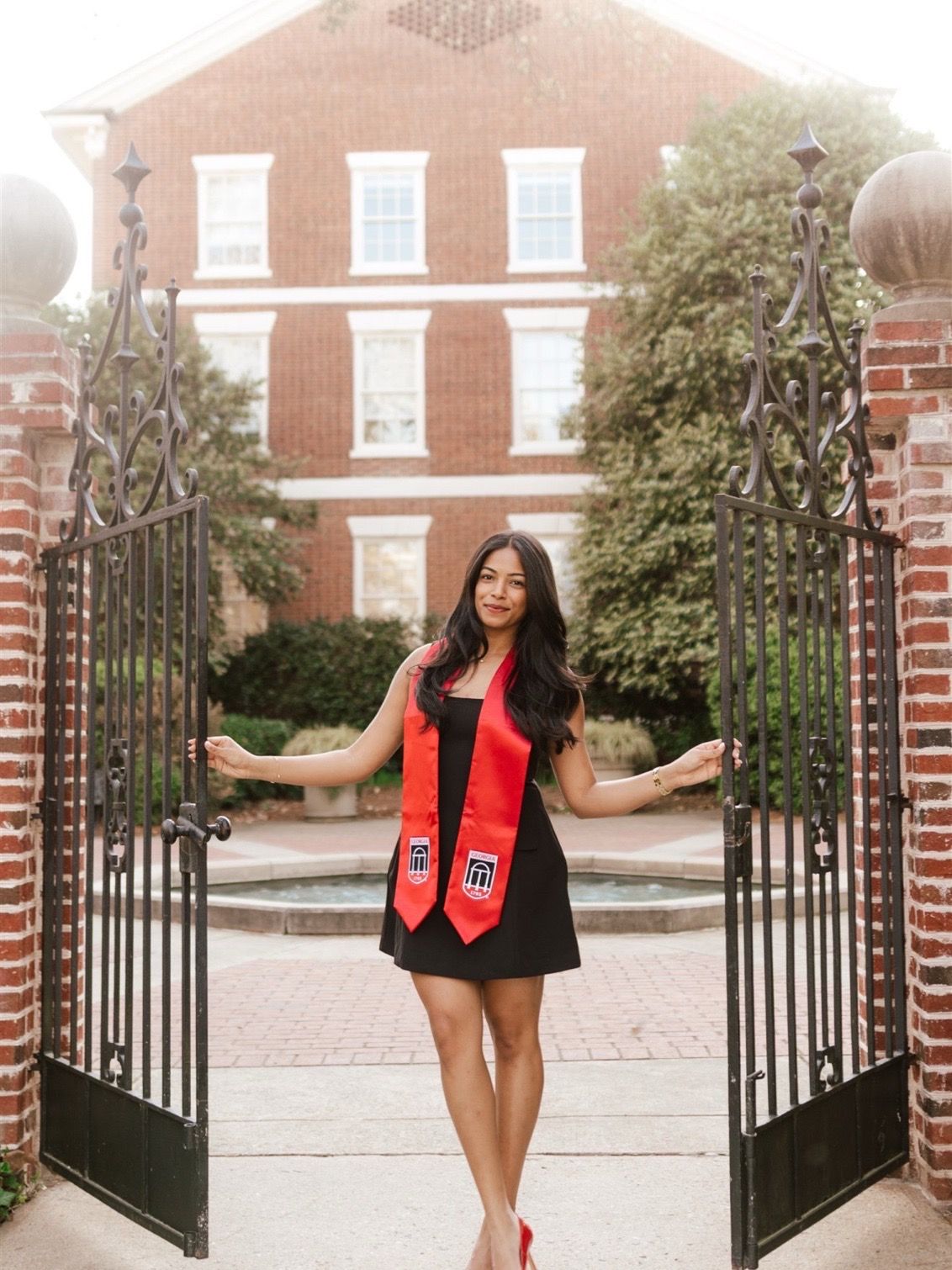 Graduate in red stole poses joyfully at ornate brick entrance with iron gates.