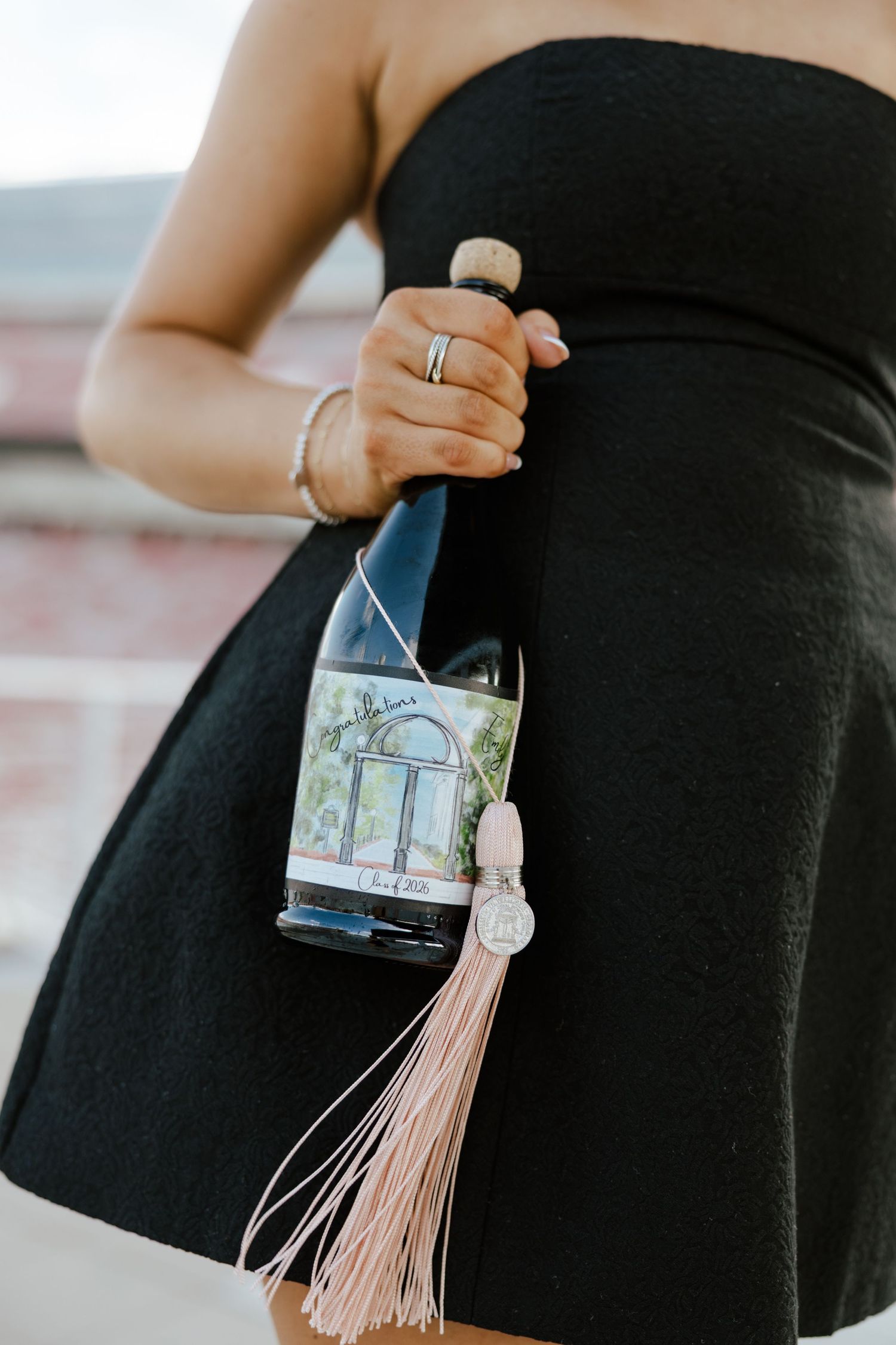 Woman in black dress holding a wine bottle with a custom label and pink tassel charm decoration.