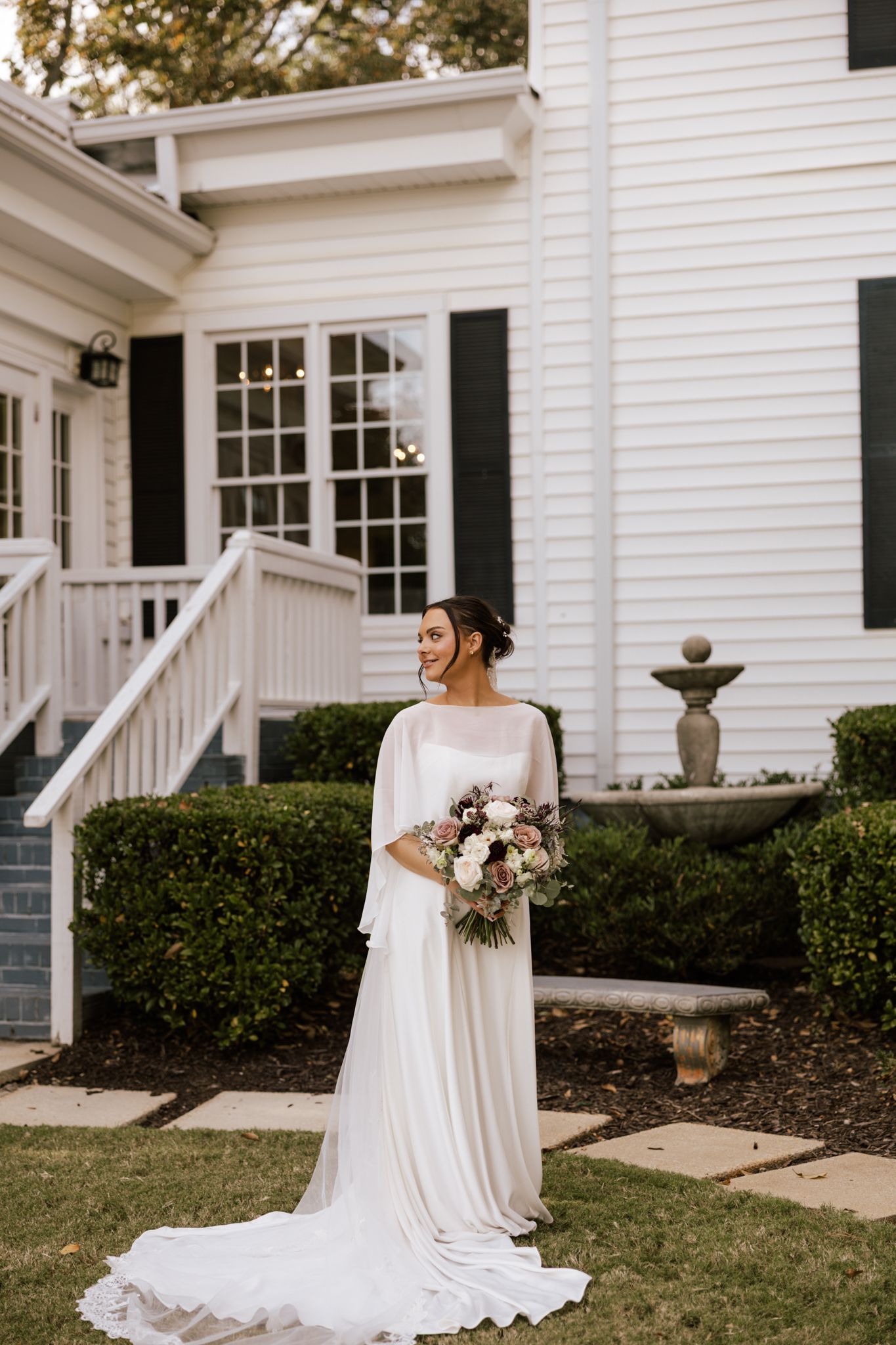 Elegant bride in flowing white cape gown holds bouquet outside a charming white colonial wedding venue.