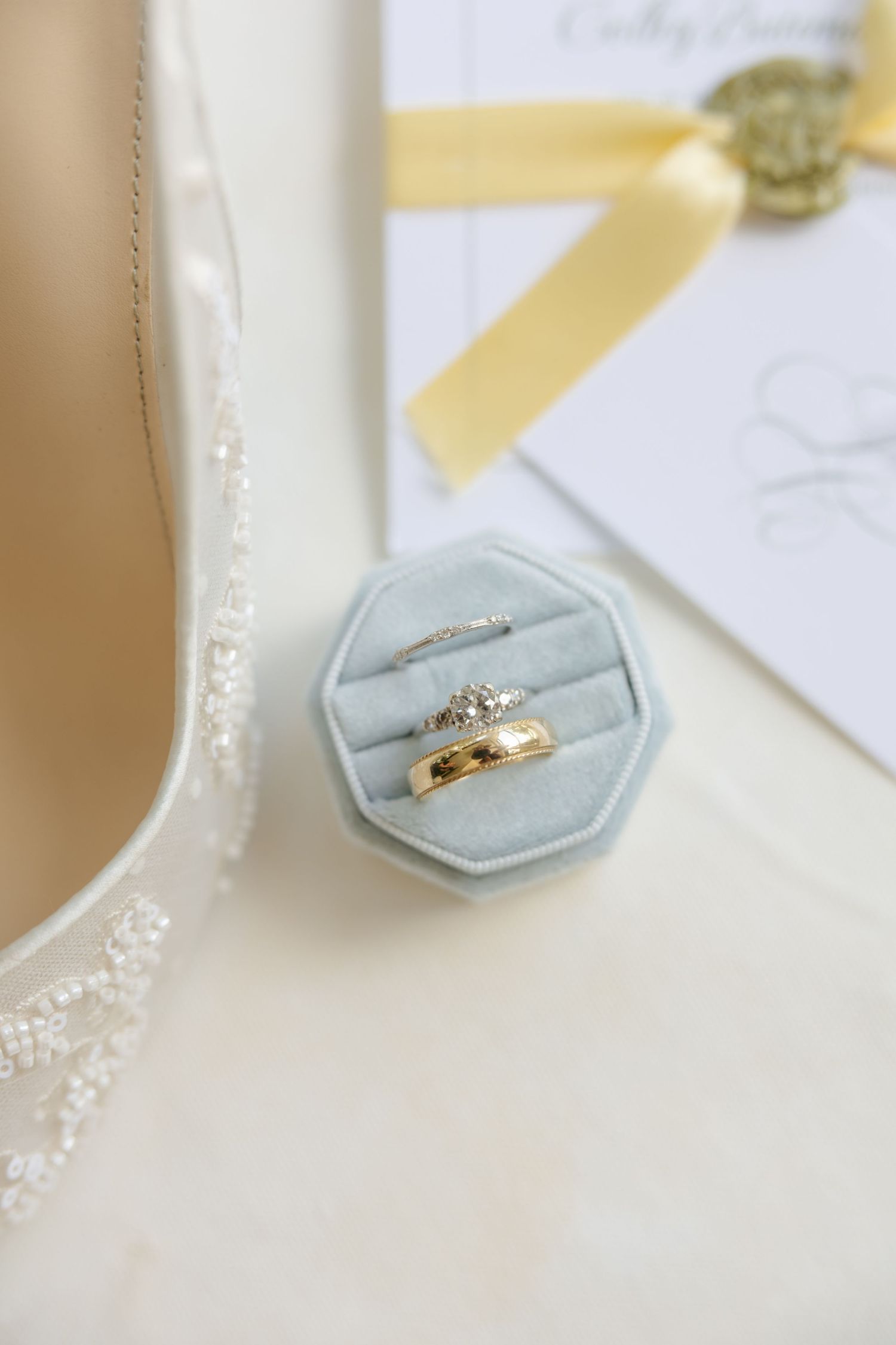 Gold wedding bands and diamond engagement ring displayed in sage velvet ring box beside bridal heels and invitation.