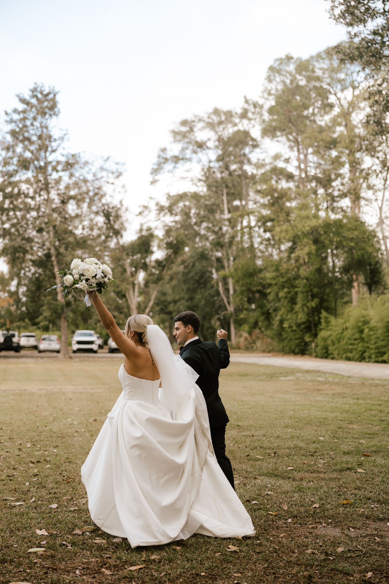 Bride and groom celebrating outdoors, bride raising bouquet of white flowers, groom in black tuxedo, lush green trees background.
