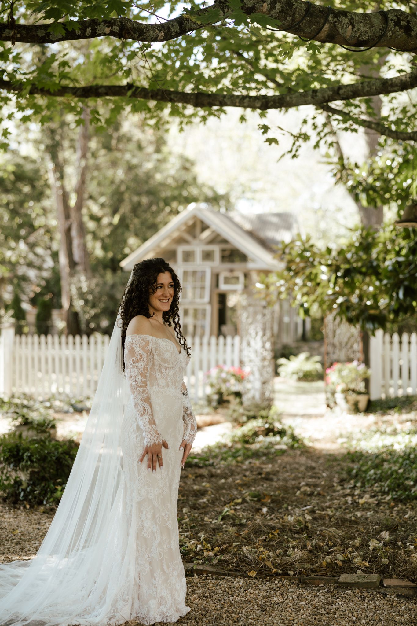 Bride in lace long-sleeve wedding gown with veil poses in garden with white picket fence and cottage backdrop.