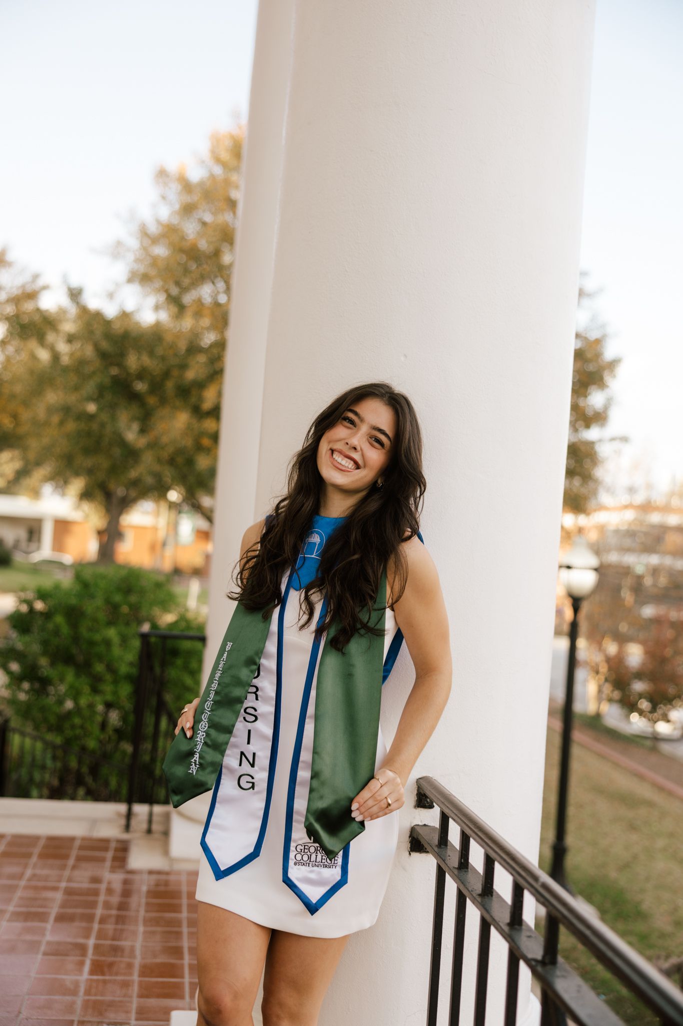 Smiling young woman in white dress wearing green and blue graduation stole poses by white column outdoors.