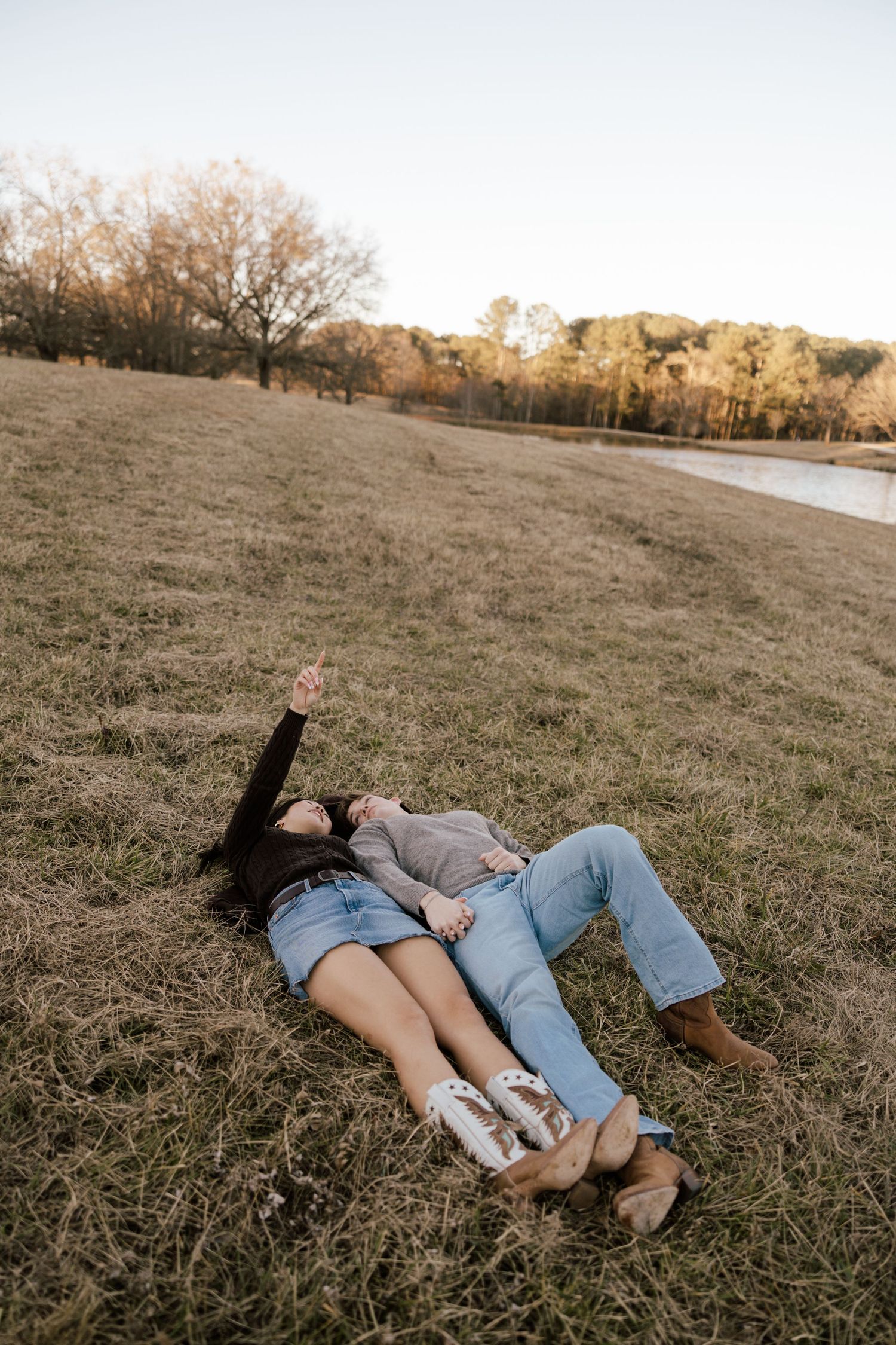 Couple laying on grass field near a pond, pointing at the sky, wearing jeans and casual tops on a fall day.
