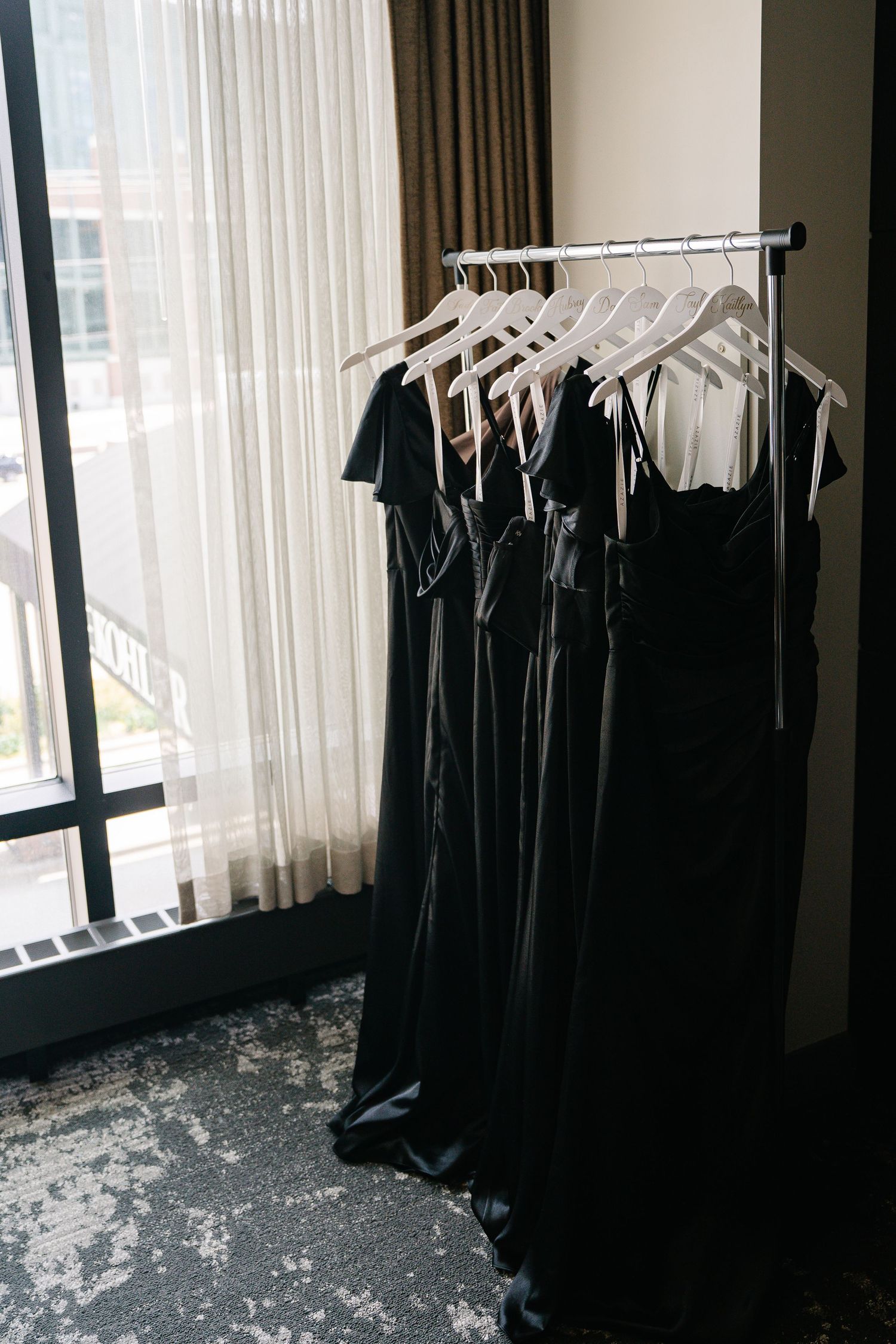 Row of black bridesmaid dresses hanging on garment rack in hotel room.