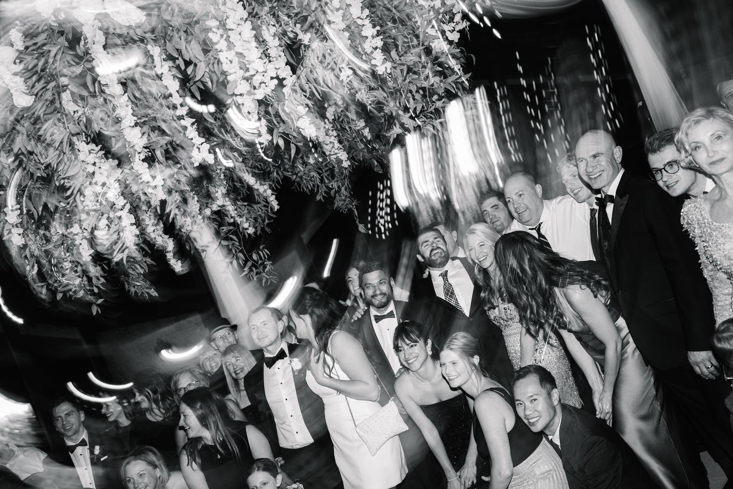 Black and white photo of people dancing and celebrating at a lively wedding reception under decorative lights.