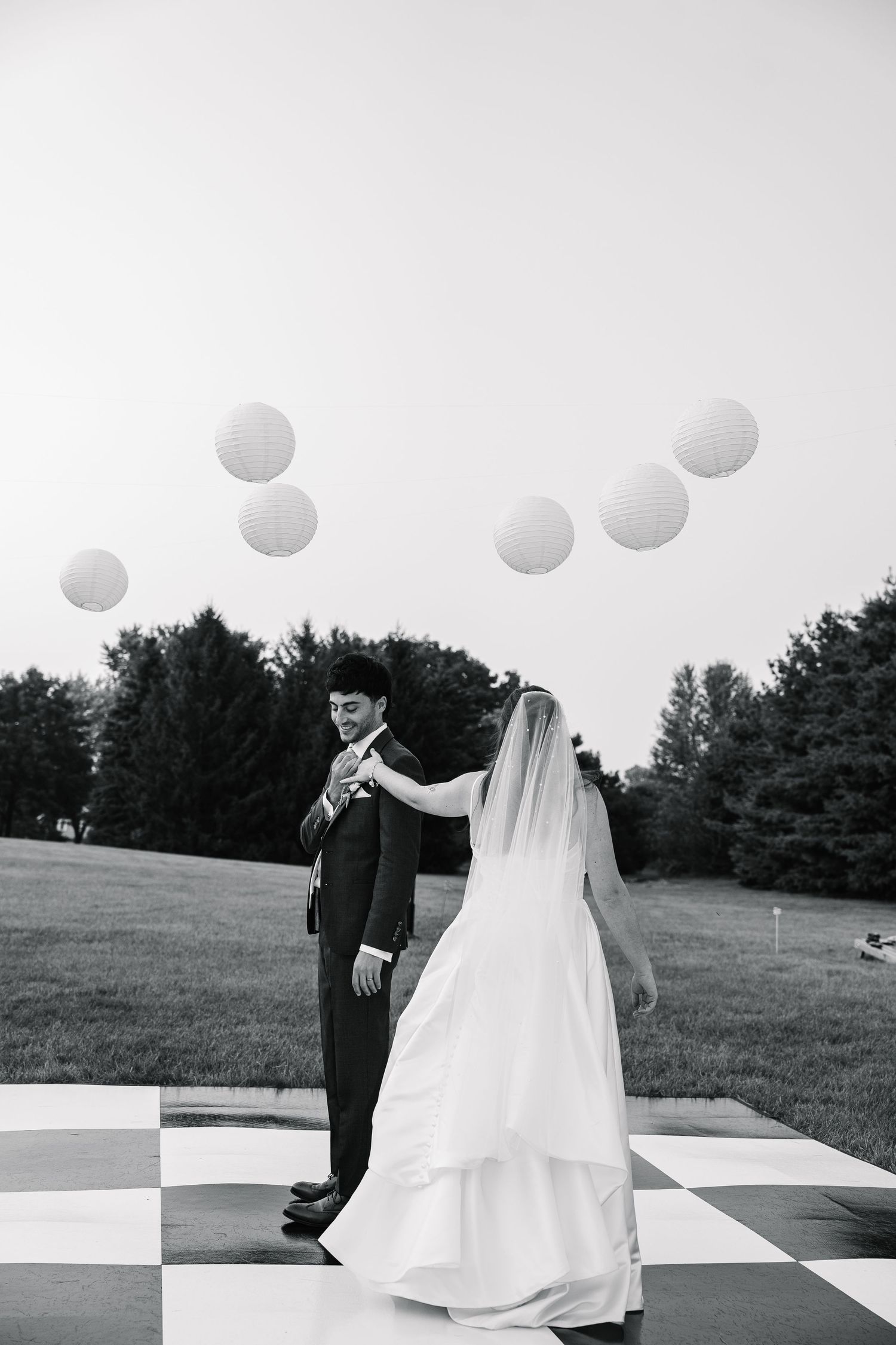 A black and white wedding photo shows a couple dancing on a giant outdoor chess board in a scenic field.
