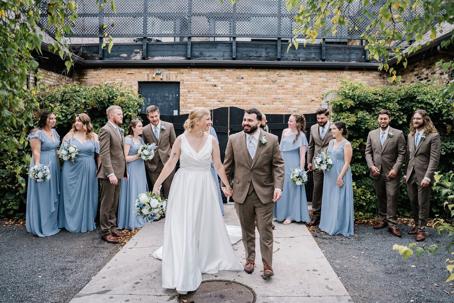 Wedding party in light blue bridesmaid dresses and tan suits poses outdoors at rustic venue with brick wall and greenery.