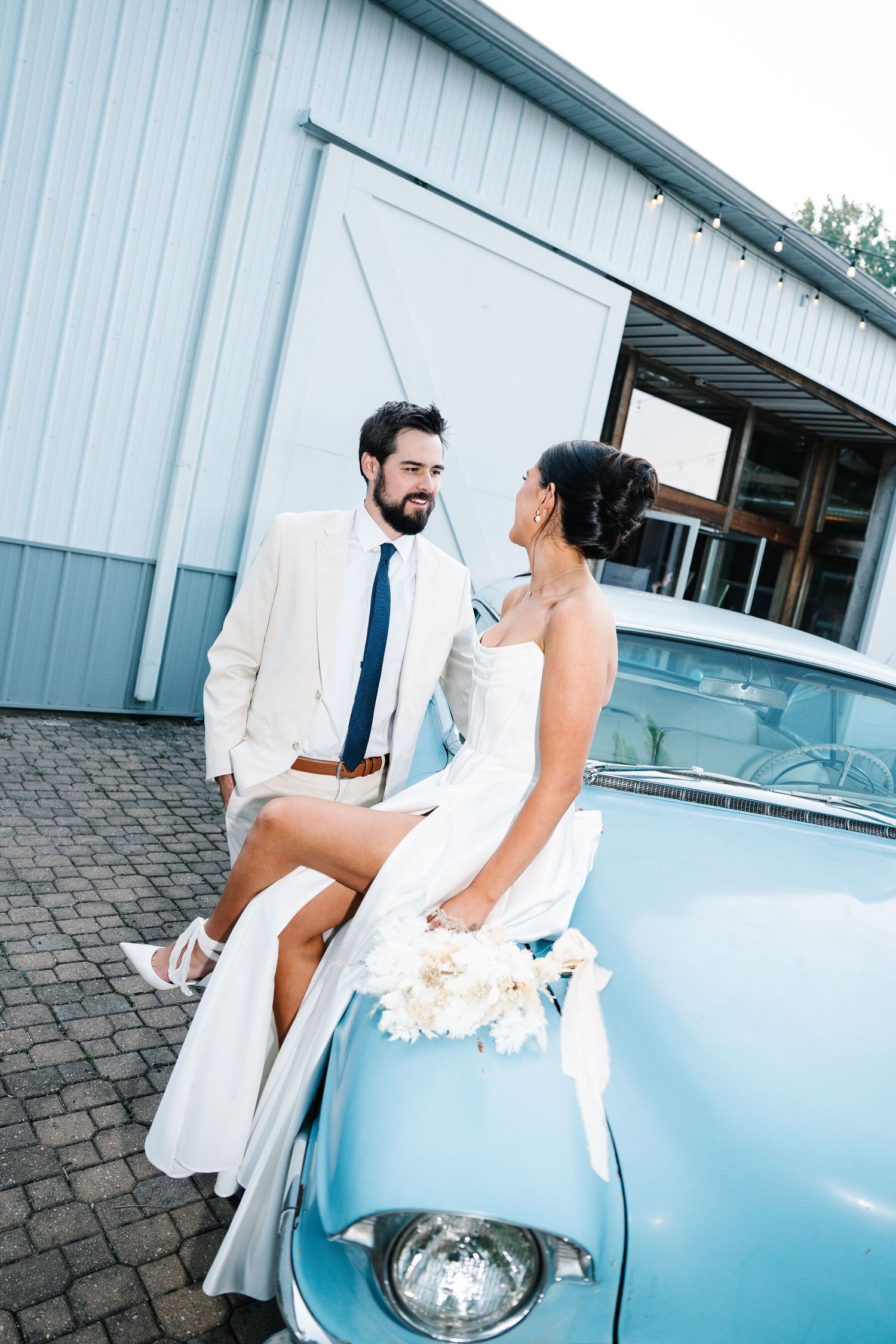 A couple in wedding attire poses together near a classic light blue vintage car.