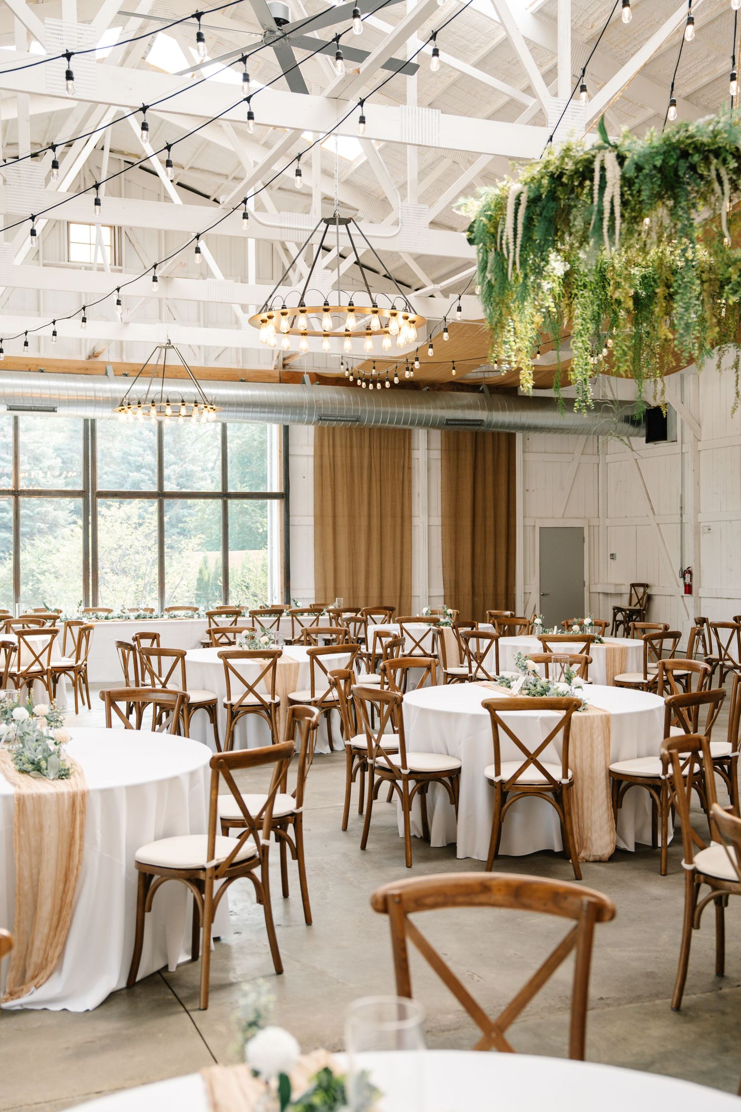 Modern reception venue with white tablecloths, wooden crossback chairs, and string lights creating an elegant atmosphere.