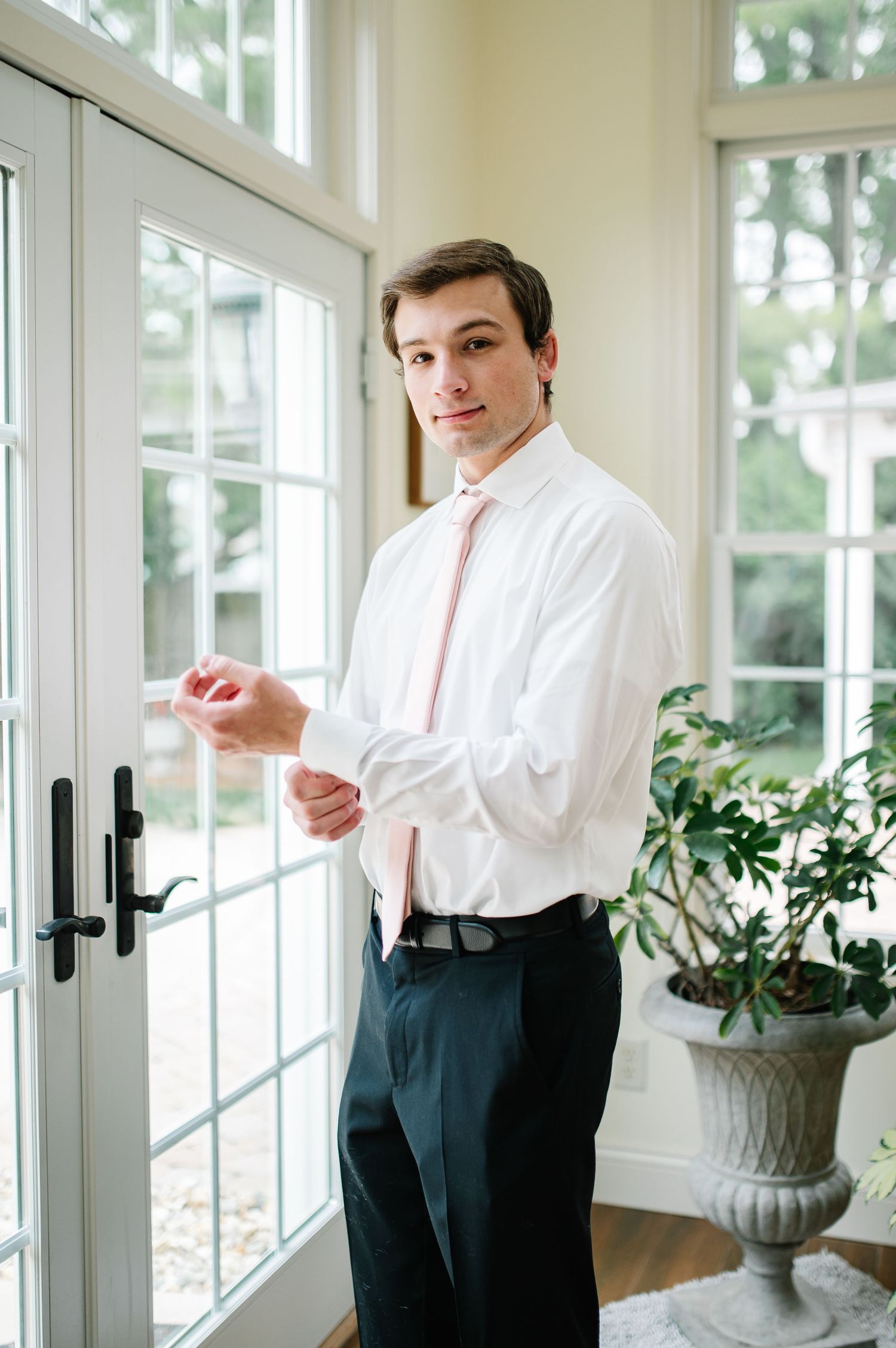 A person in formal attire adjusts their clothing while standing in a bright sunlit room with French doors and potted plants.