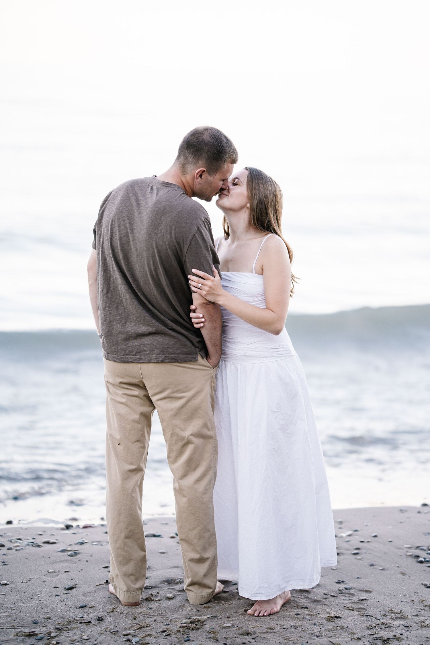 Couple sharing a romantic kiss on a beach, woman in white dress, man in gray shirt and khaki pants, ocean waves behind them.