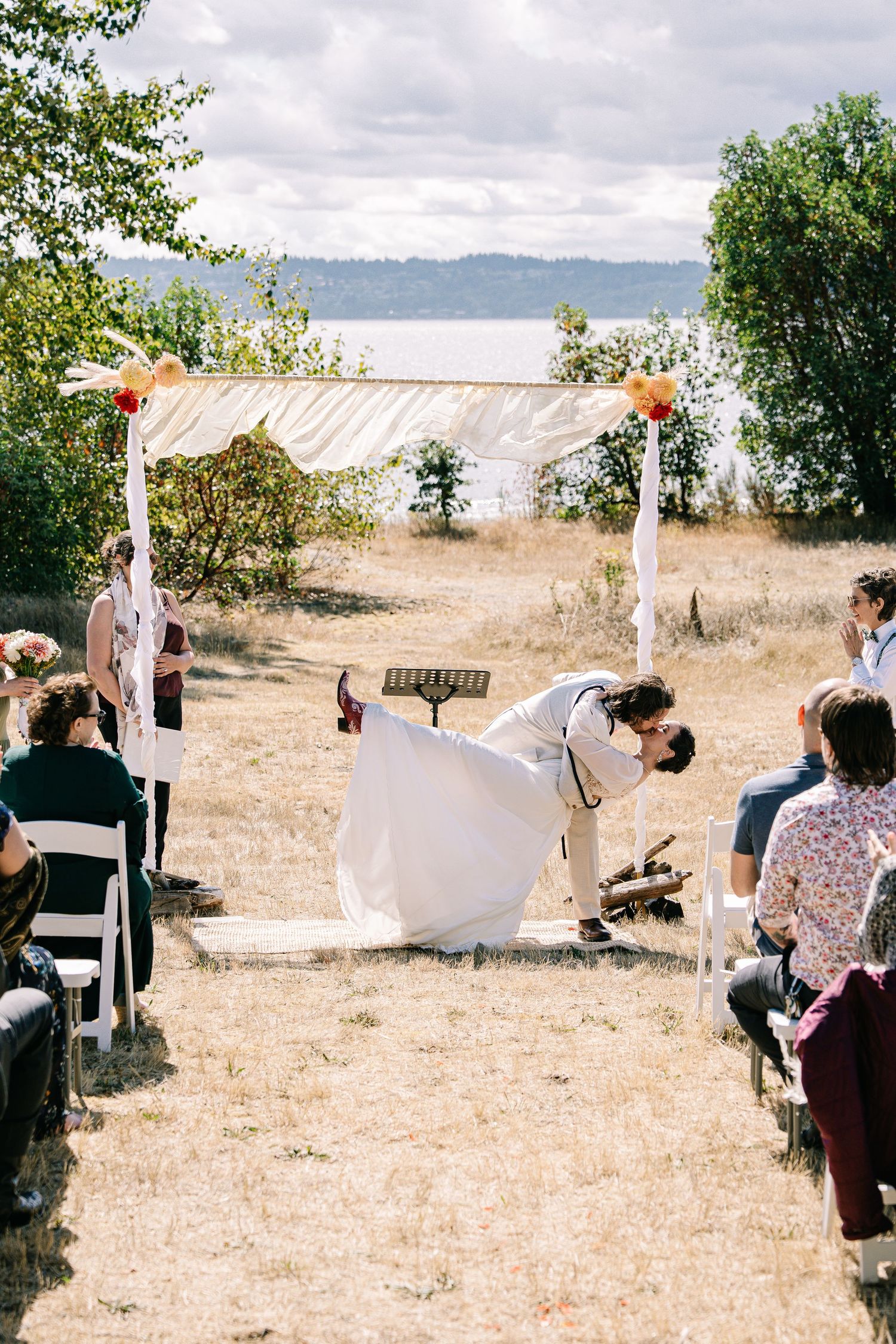 A rustic outdoor wedding ceremony with white fabric draped arbor and chairs set up in a natural field setting.