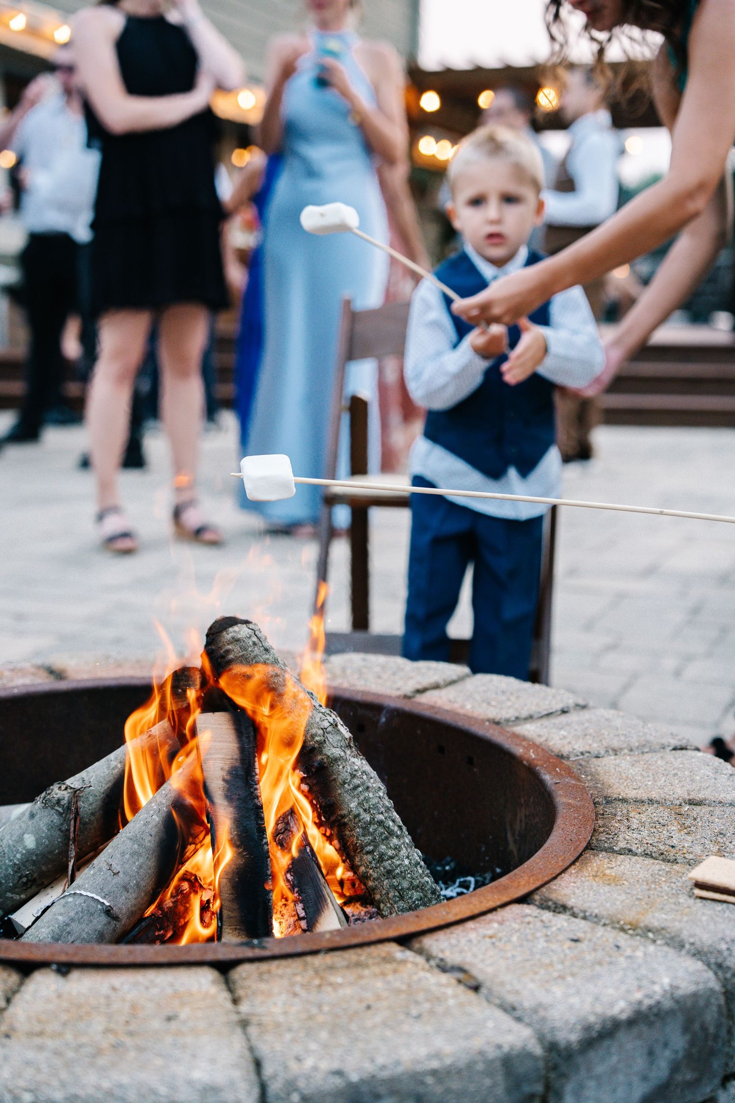A rustic outdoor fire pit with glowing flames surrounded by guests at an evening gathering.
