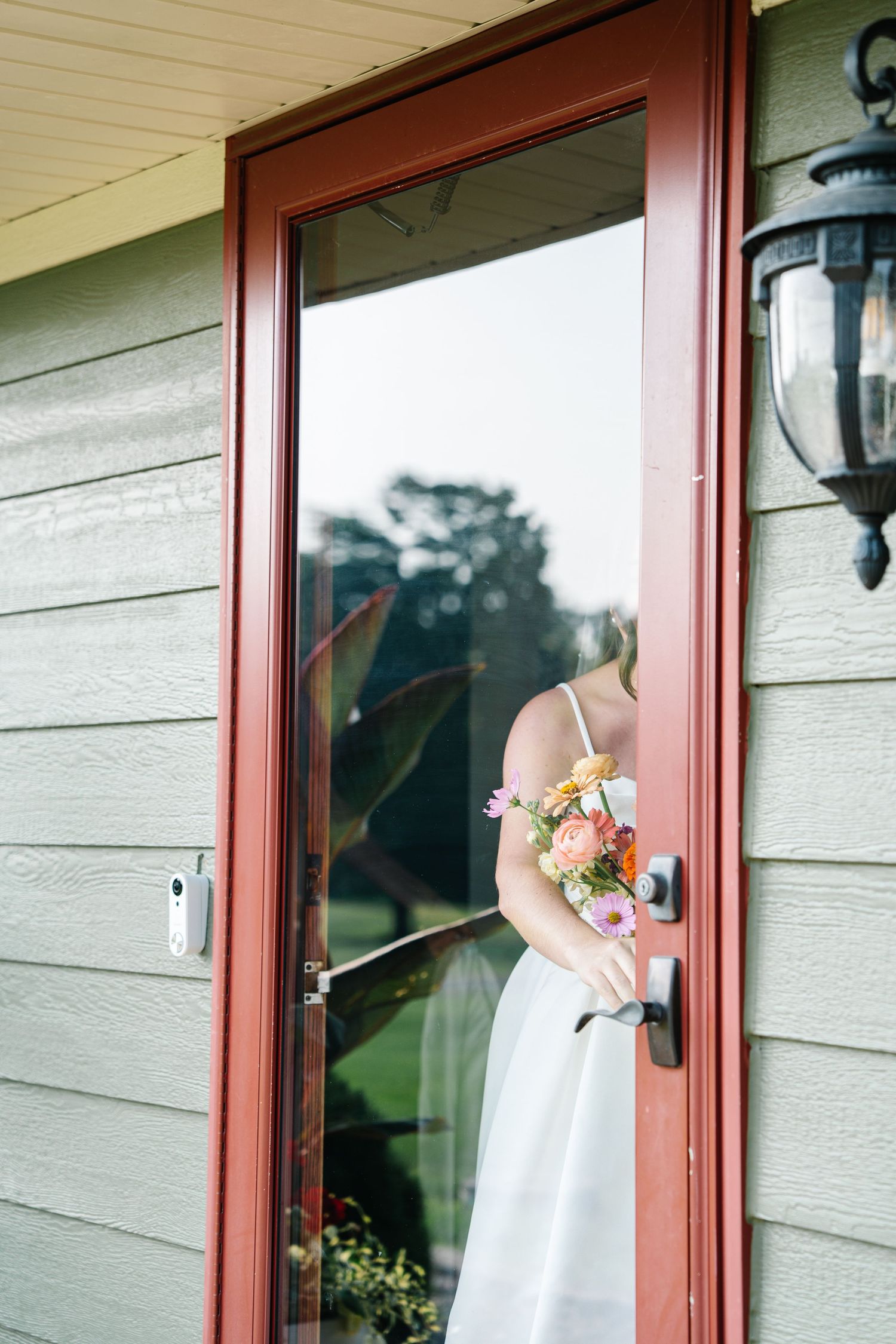 A white wedding dress and floral bouquet are visible through a red doorframe of a house with a wall lantern.