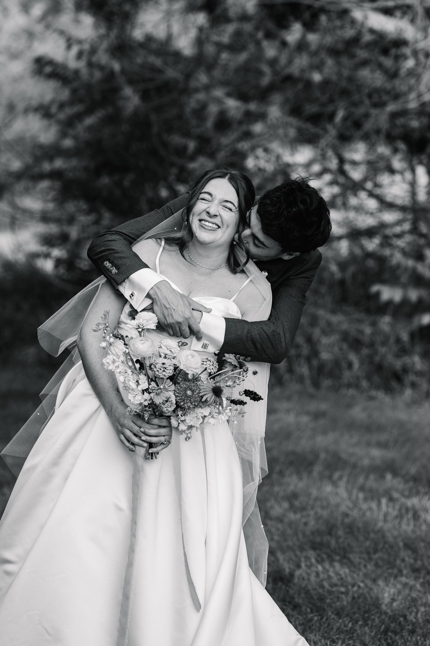 Two people in white dresses share joyful candid moments together outdoors in a black and white photograph series.