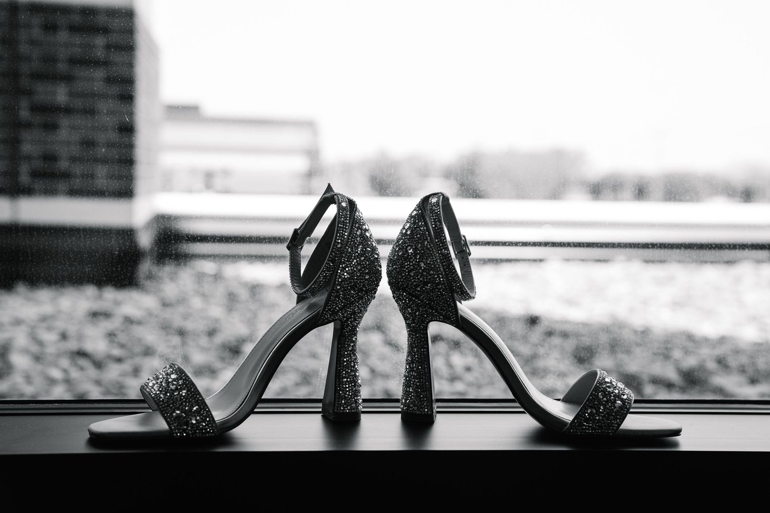 A pair of sparkly black high heel shoes sits on a windowsill in black and white photography.