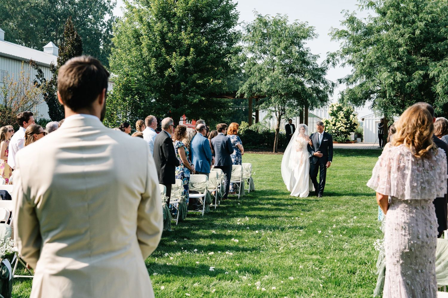 Outdoor wedding ceremony with guests standing in rows on a grassy lawn surrounded by trees on a sunny day.
