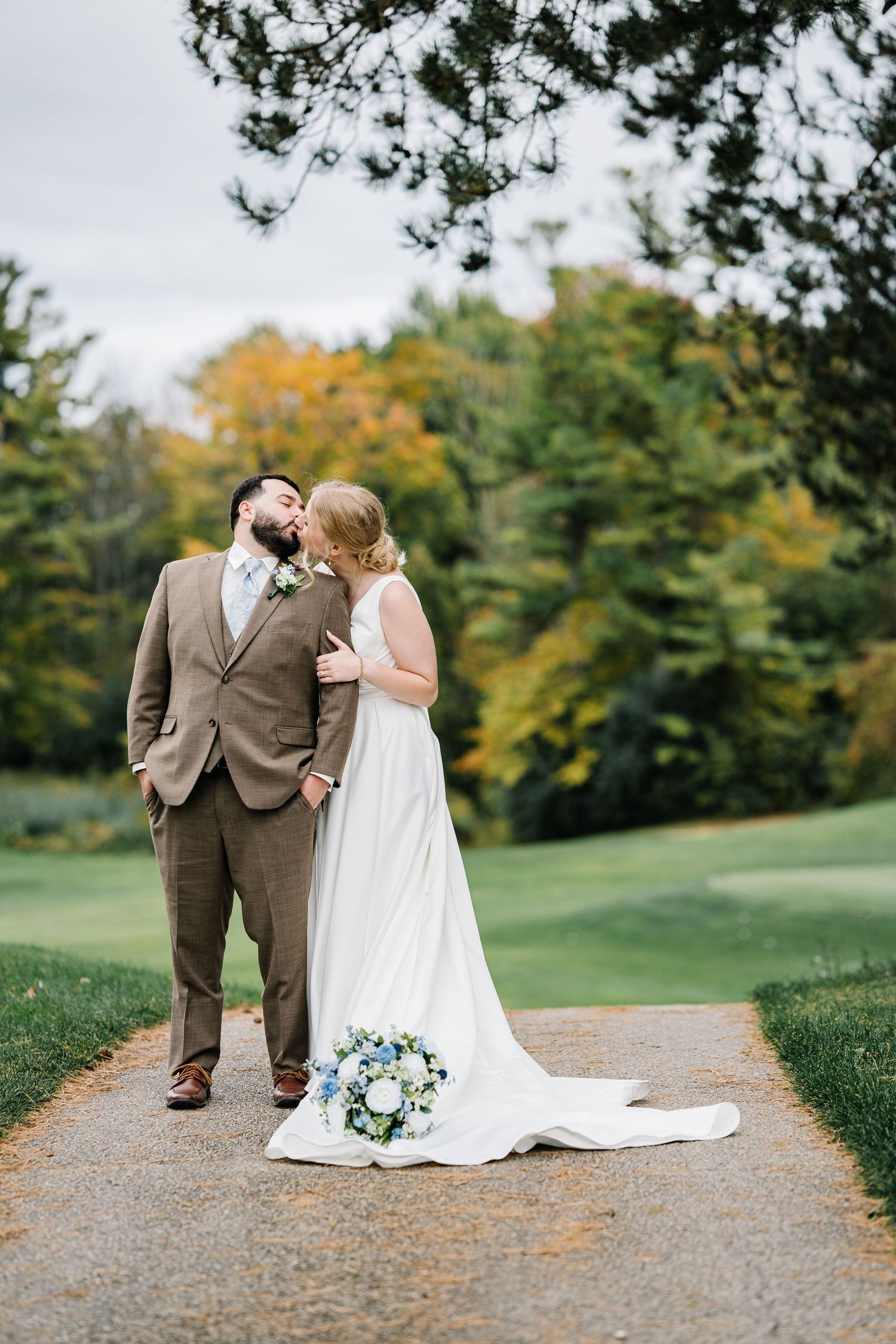 A couple in wedding attire shares an intimate moment while walking on a gravel path surrounded by autumn foliage.