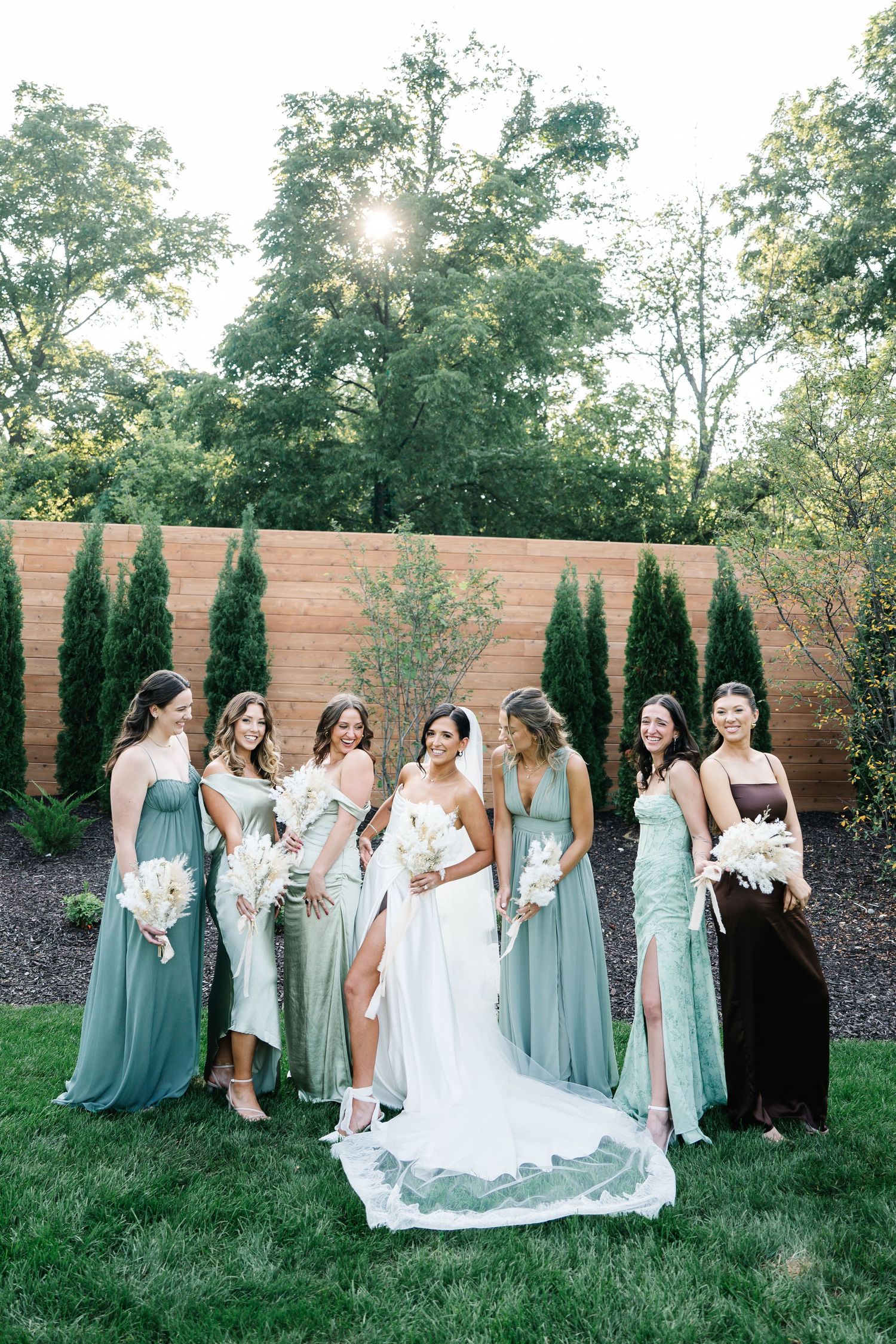 A wedding party in seafoam green bridesmaid dresses poses together on a lawn with a wooden fence backdrop.