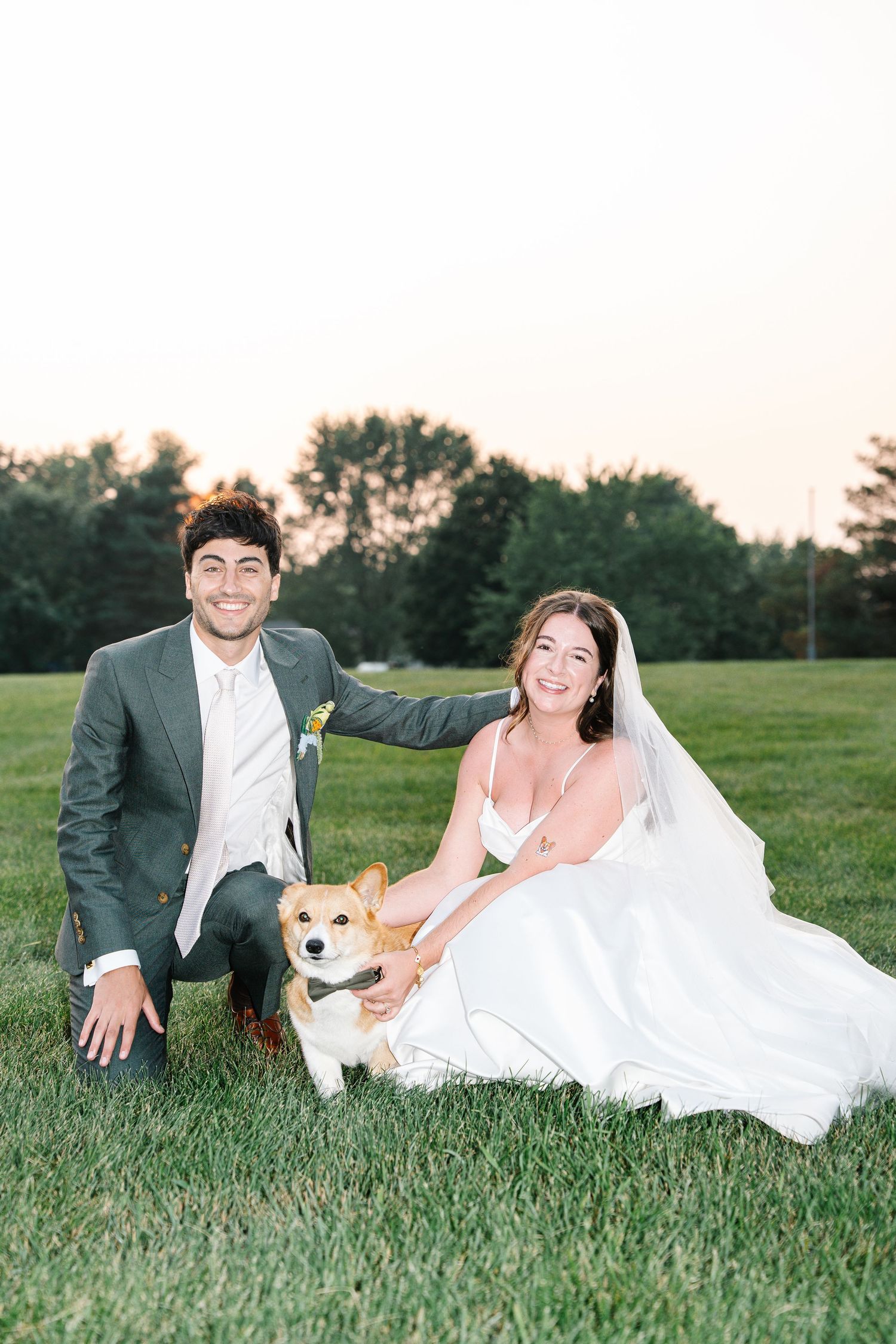 A couple in wedding attire laying in green grass with their white dog during an outdoor photo session.