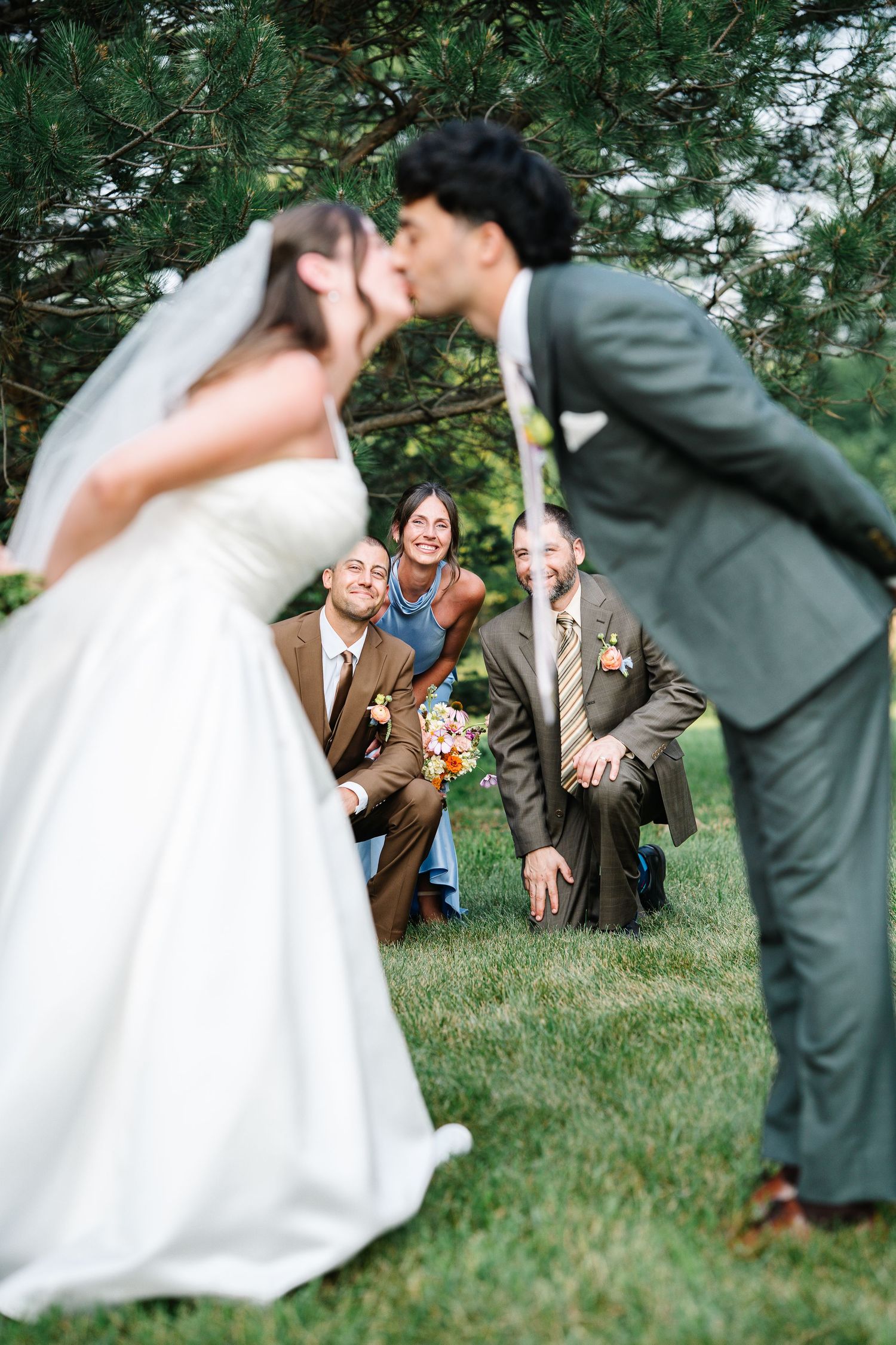 A playful wedding couple dips and poses in a series of romantic shots on a grassy lawn with trees in the background.