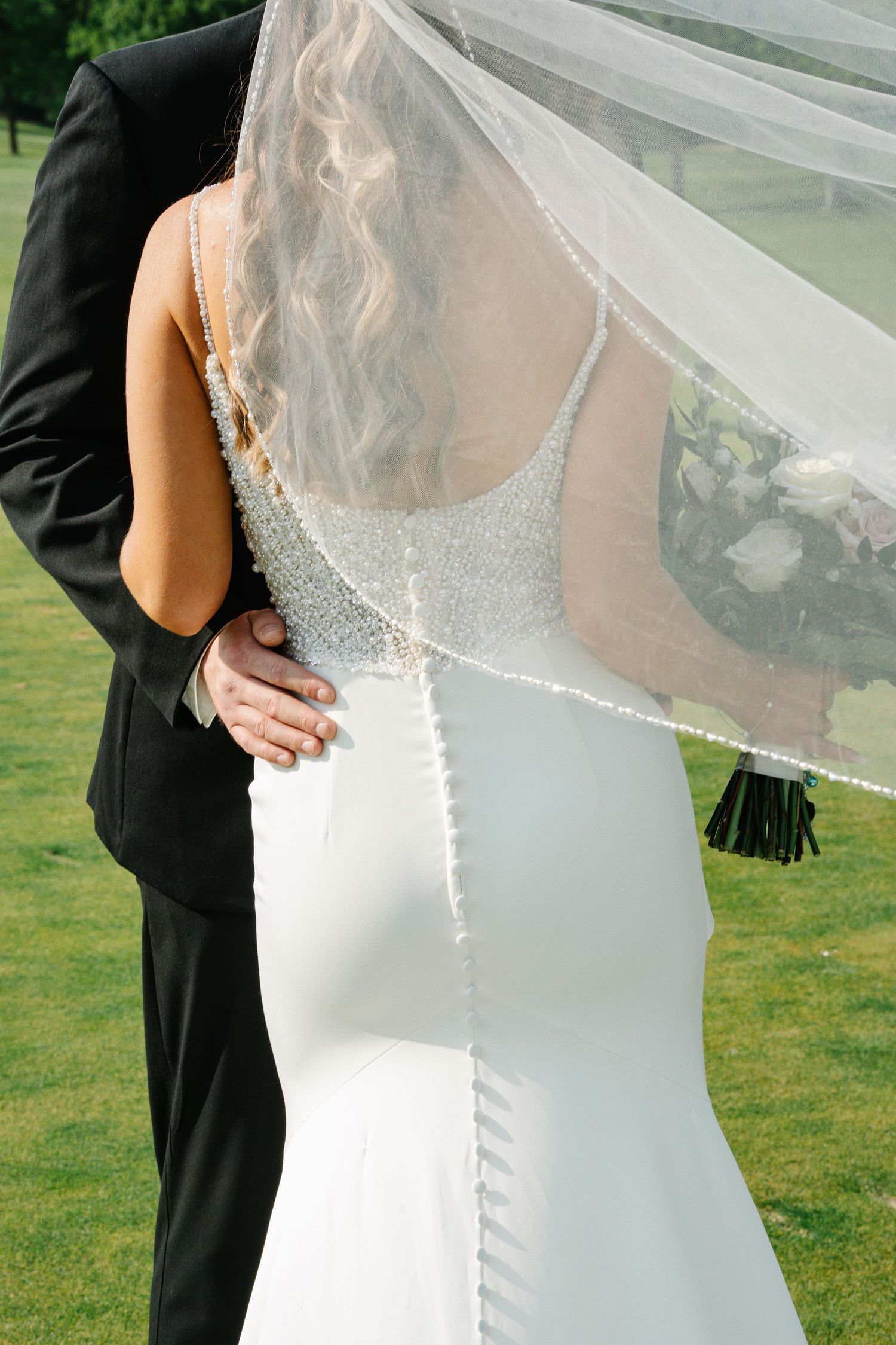 A bride in a white beaded wedding dress stands with her groom on a grassy lawn under a delicate veil.