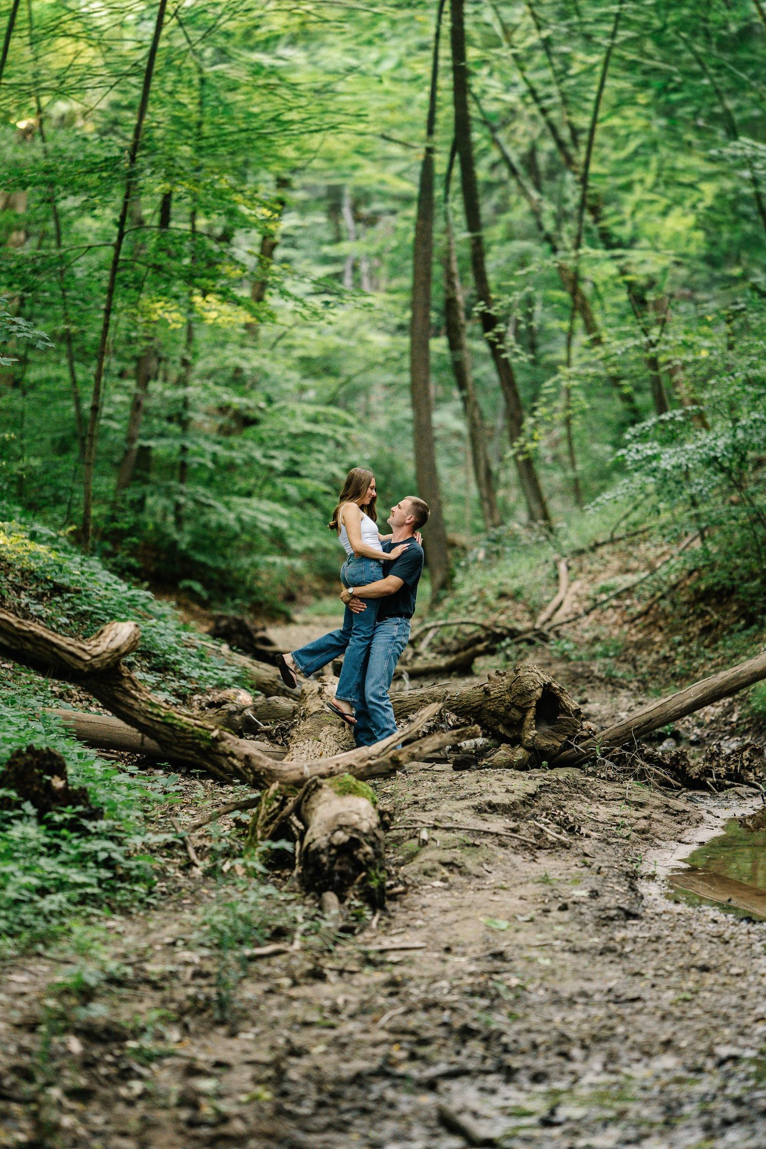 Couple embracing romantically on fallen logs in a lush green forest with muddy ground and tall trees.