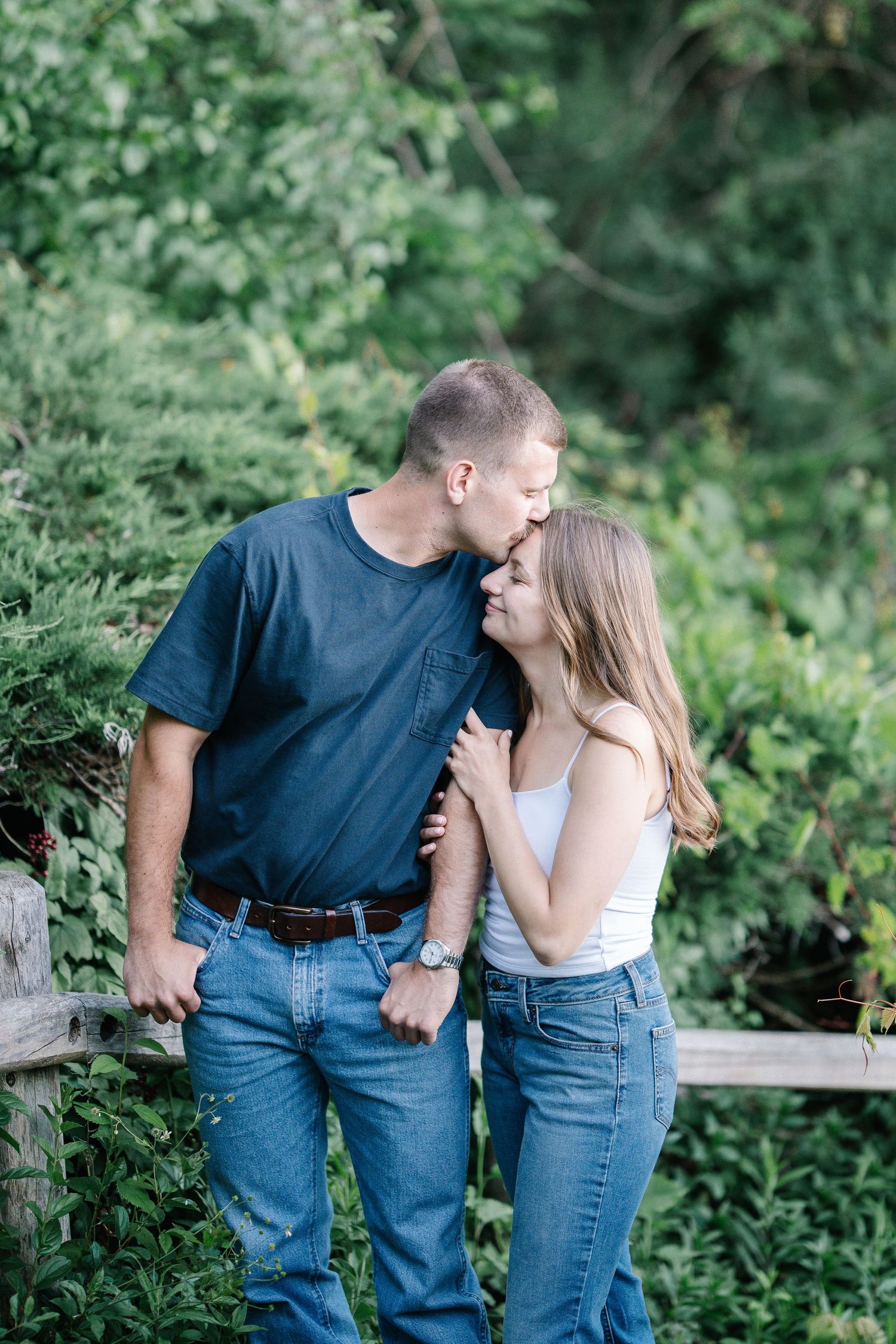 A couple shares a tender moment outdoors, man kissing woman's forehead, both wearing jeans near a wooden fence with lush greenery.
