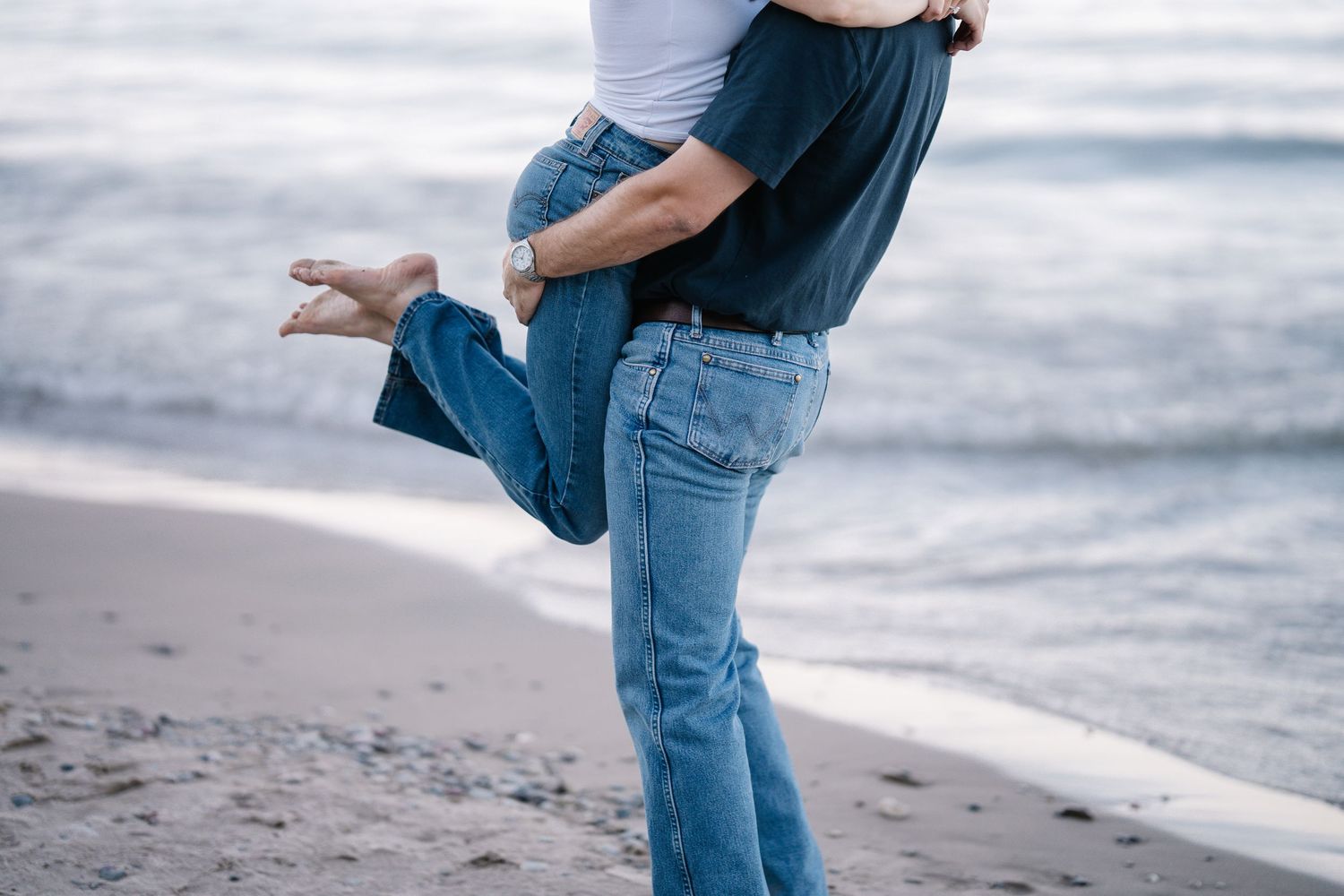 Couple embracing on a sandy beach, man lifting woman in jeans near ocean waves.