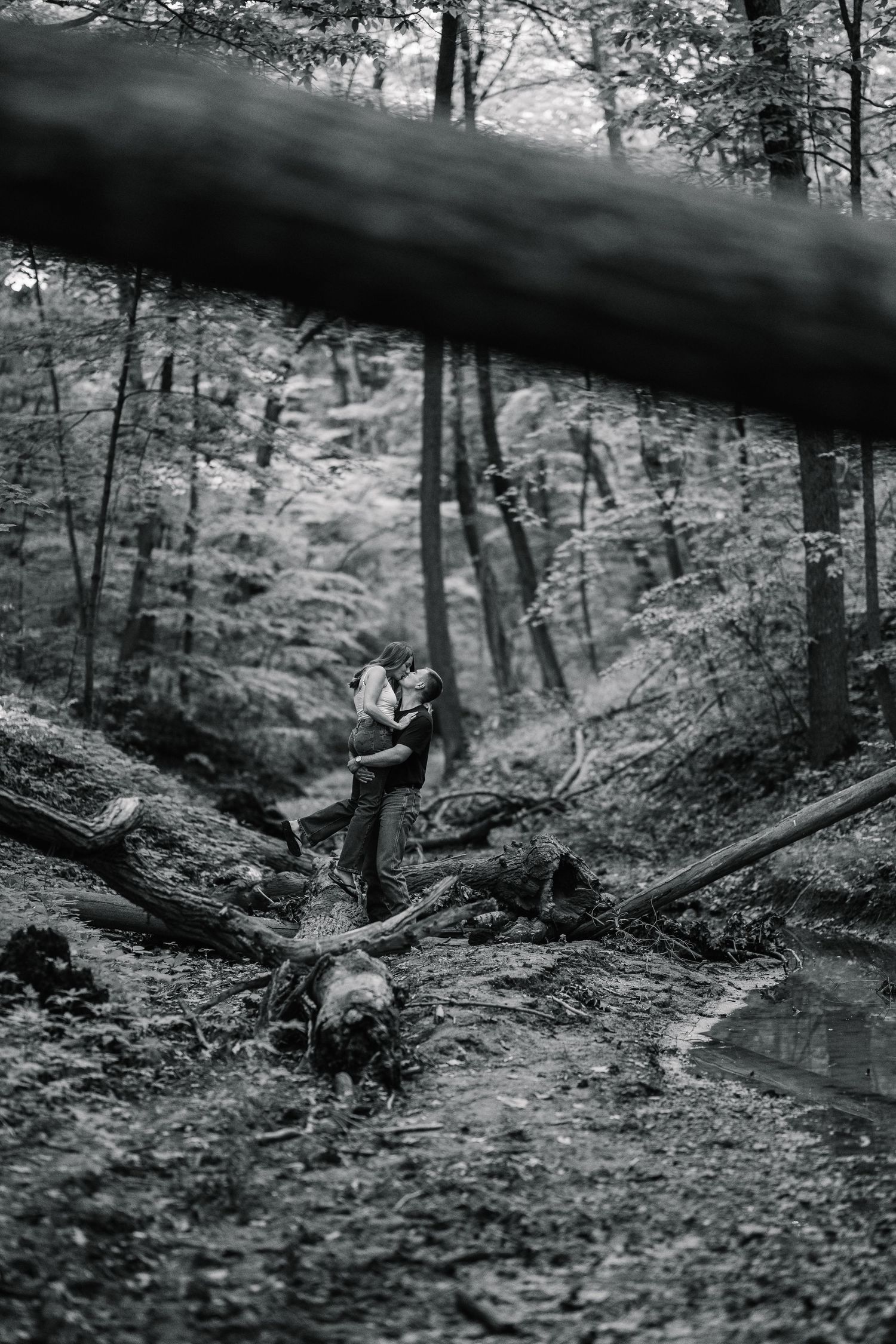 Black and white photo of a couple embracing in a lush forest surrounded by fallen logs and trees.
