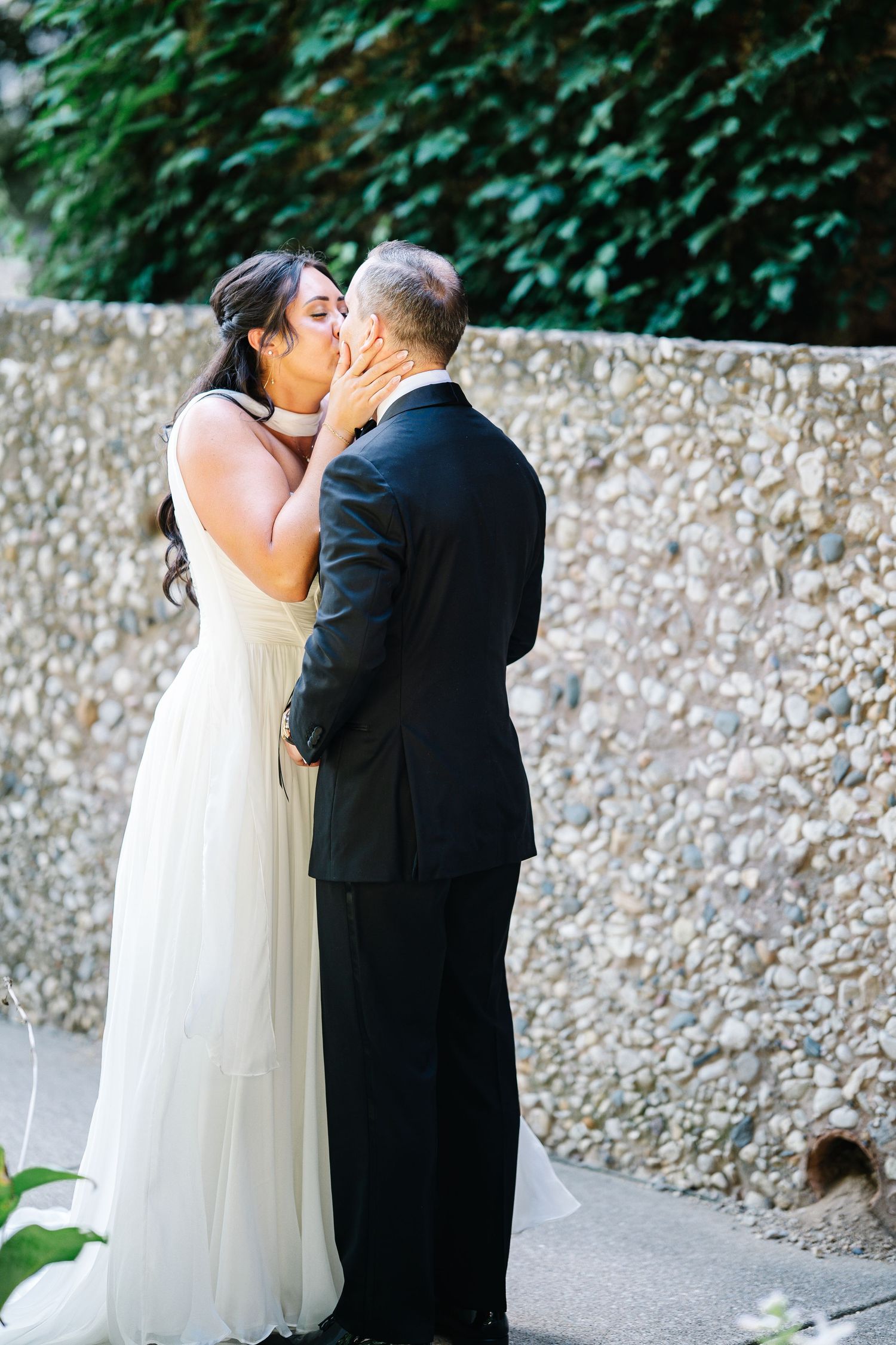 A romantic wedding moment with a couple sharing a kiss against a stone wall backdrop in an outdoor garden setting.