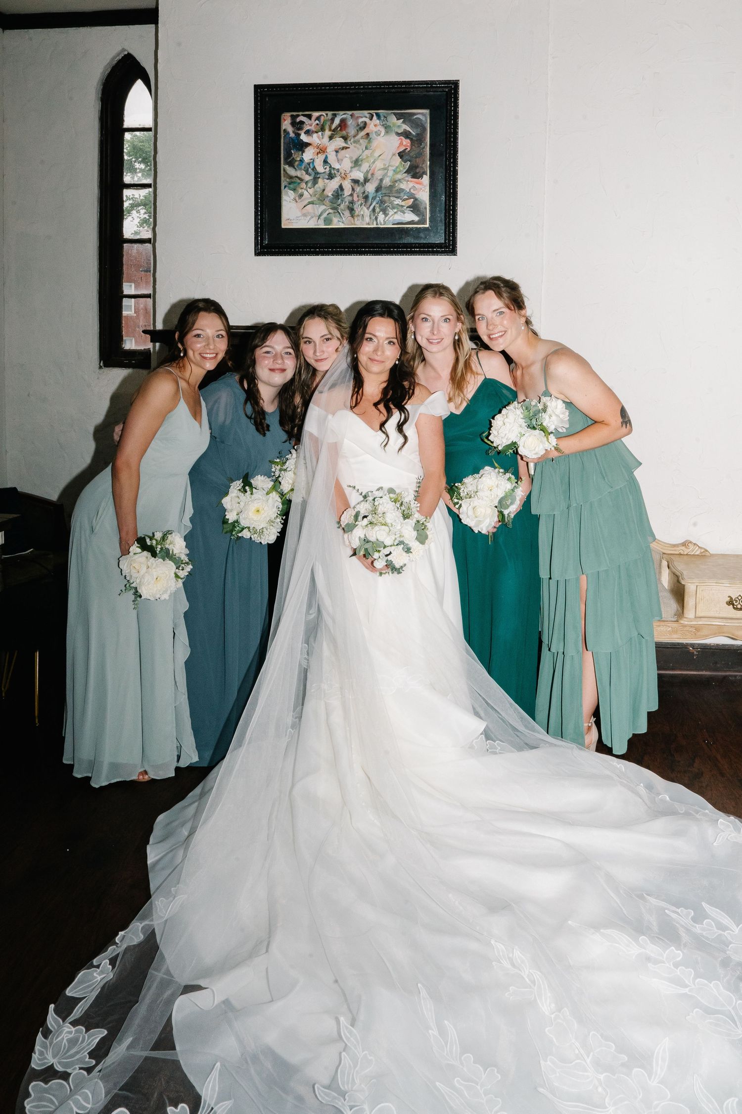 Wedding party poses in coordinated green bridesmaid dresses alongside bride in flowing white gown with long train.