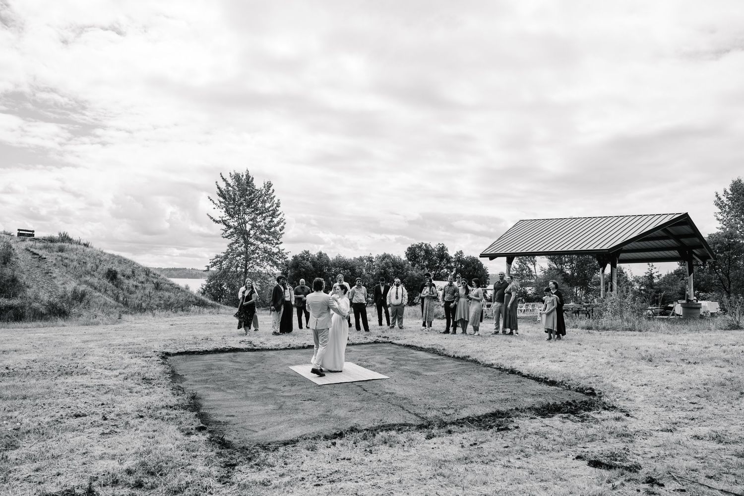 Black and white photo of outdoor wedding ceremony setup with guests gathering under a rustic pavilion.