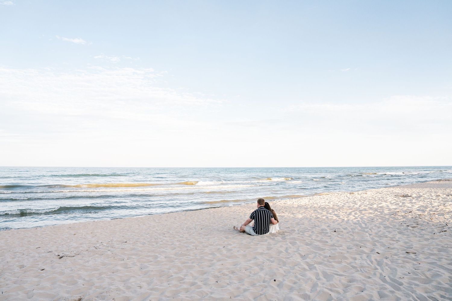 A lone person sits on a sandy beach gazing at the calm ocean waves under a soft blue sky.