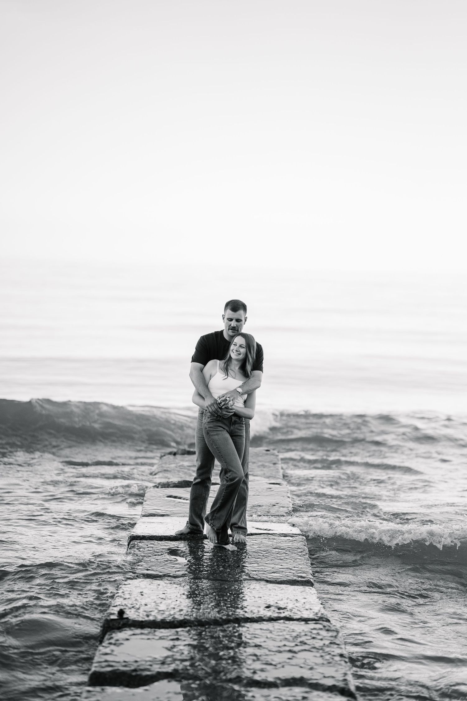 Black and white photo of a couple embracing on a wet stone pier with ocean waves in the background.