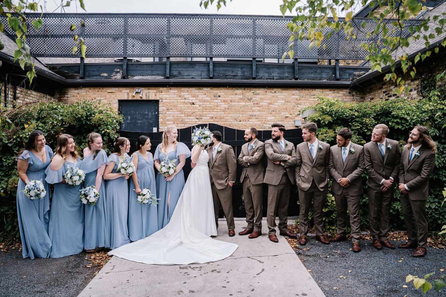 Wedding party members in light blue dresses and brown suits gather together outdoors for formal group photos.