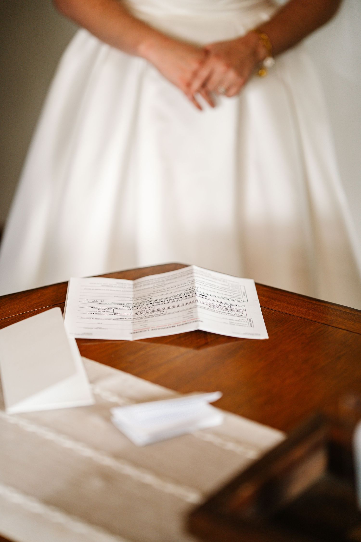 A folded paper document rests on a dark wooden table with a white wedding dress visible in the background.