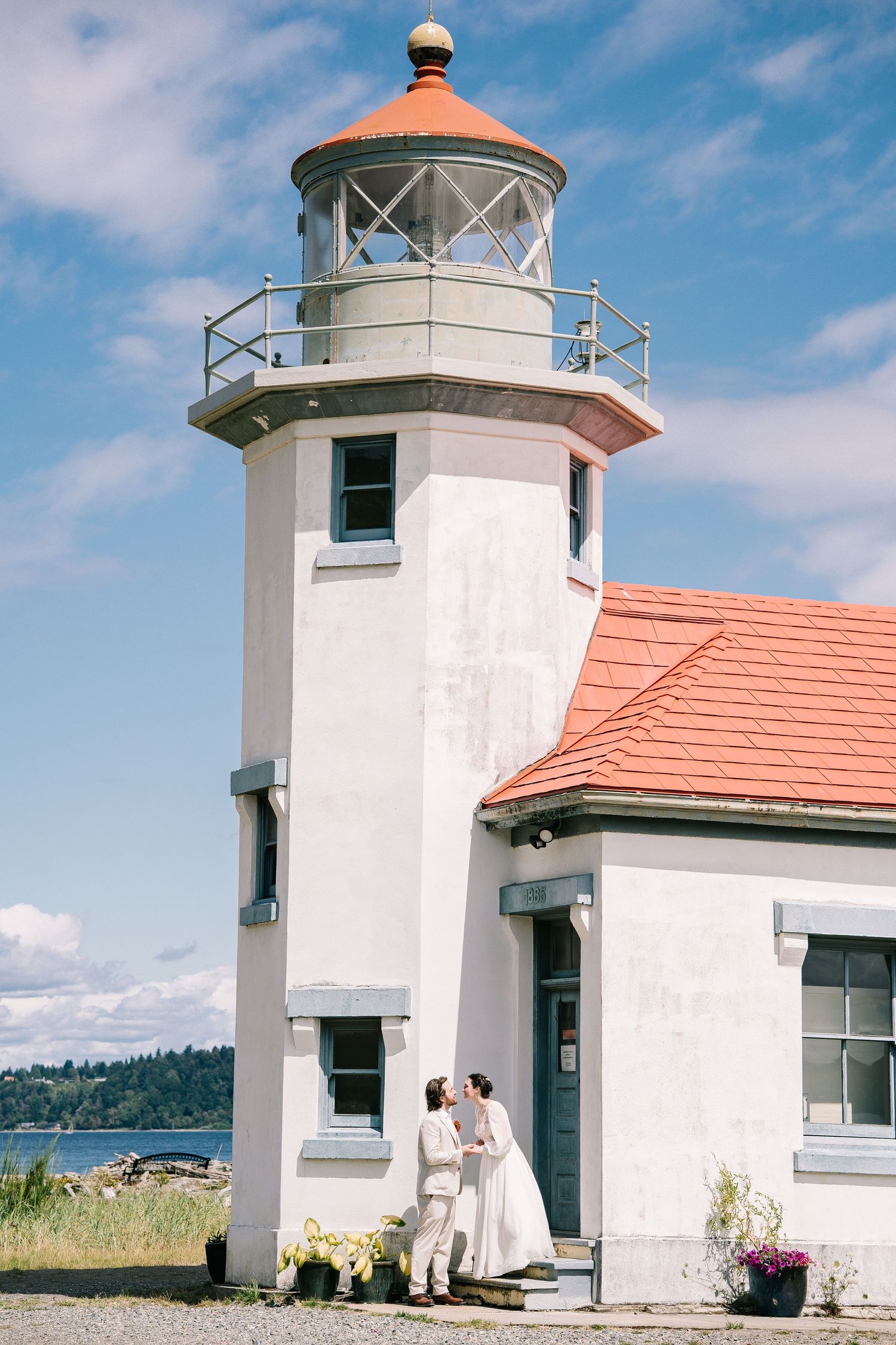 White lighthouse with orange roof and multiple towers against blue sky at waterfront location.