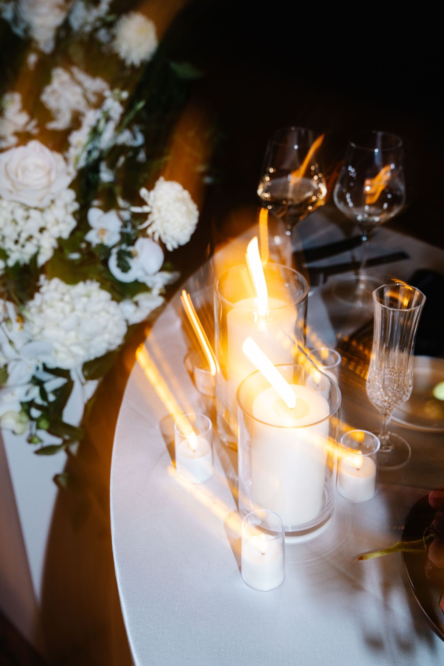 White candles with glowing flames illuminate a dark table setting with elegant floral arrangements and crystal glassware.
